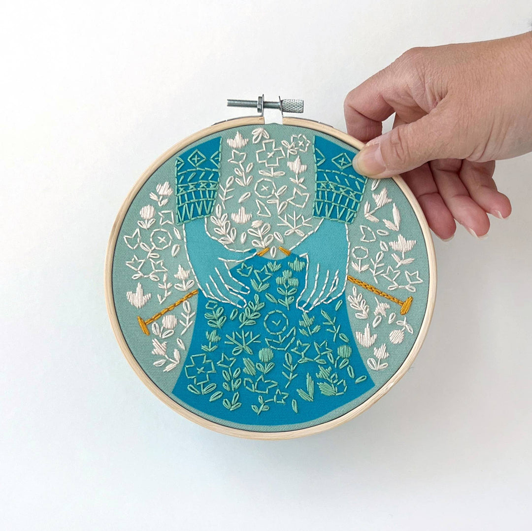 Rikrack Knit Embroidery Kit featuring illustrated hands knitting with golden needles, a patterned blue sweater, and delicate floral details in a six-inch embroidery hoop.
 Copy Alt Text