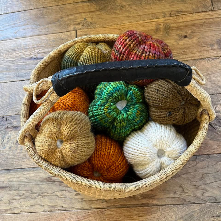 A woven basket filled with knitted pumpkins in multiple colors, including green, orange, cream, brown, and red, set on a wooden floor.