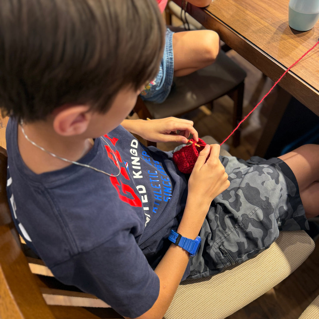 A boy in a navy blue graphic t-shirt and gray camouflage shorts sits at a wooden table, knitting a red item with red yarn. Another child is partially visible in the background.