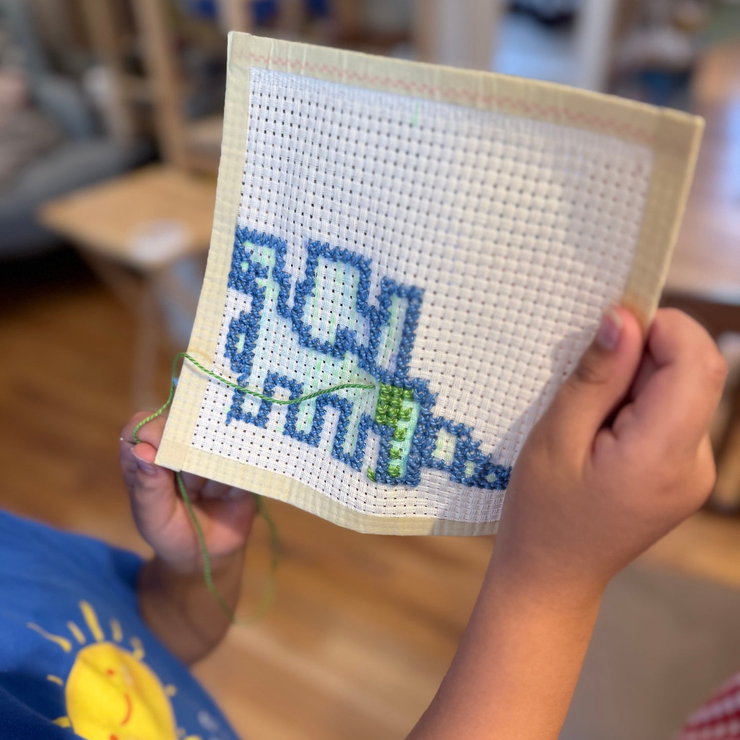 A child holds up a square piece of cross-stitch embroidery showing a blue dinosaur design on white fabric, with hands and part of a blue shirt visible in a softly blurred indoor setting.