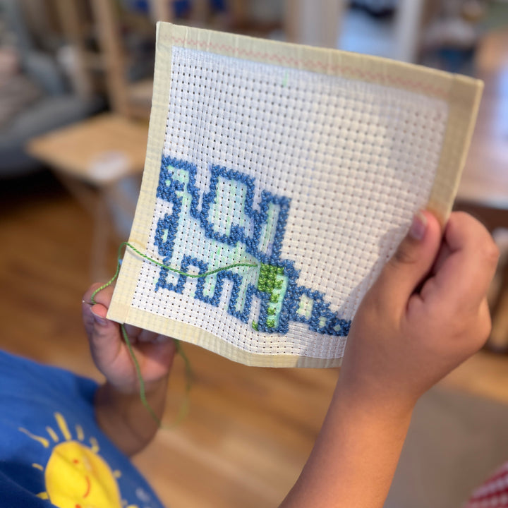 A child holds up a square piece of cross-stitch embroidery showing a blue dinosaur design on white fabric, with hands and part of a blue shirt visible in a softly blurred indoor setting.