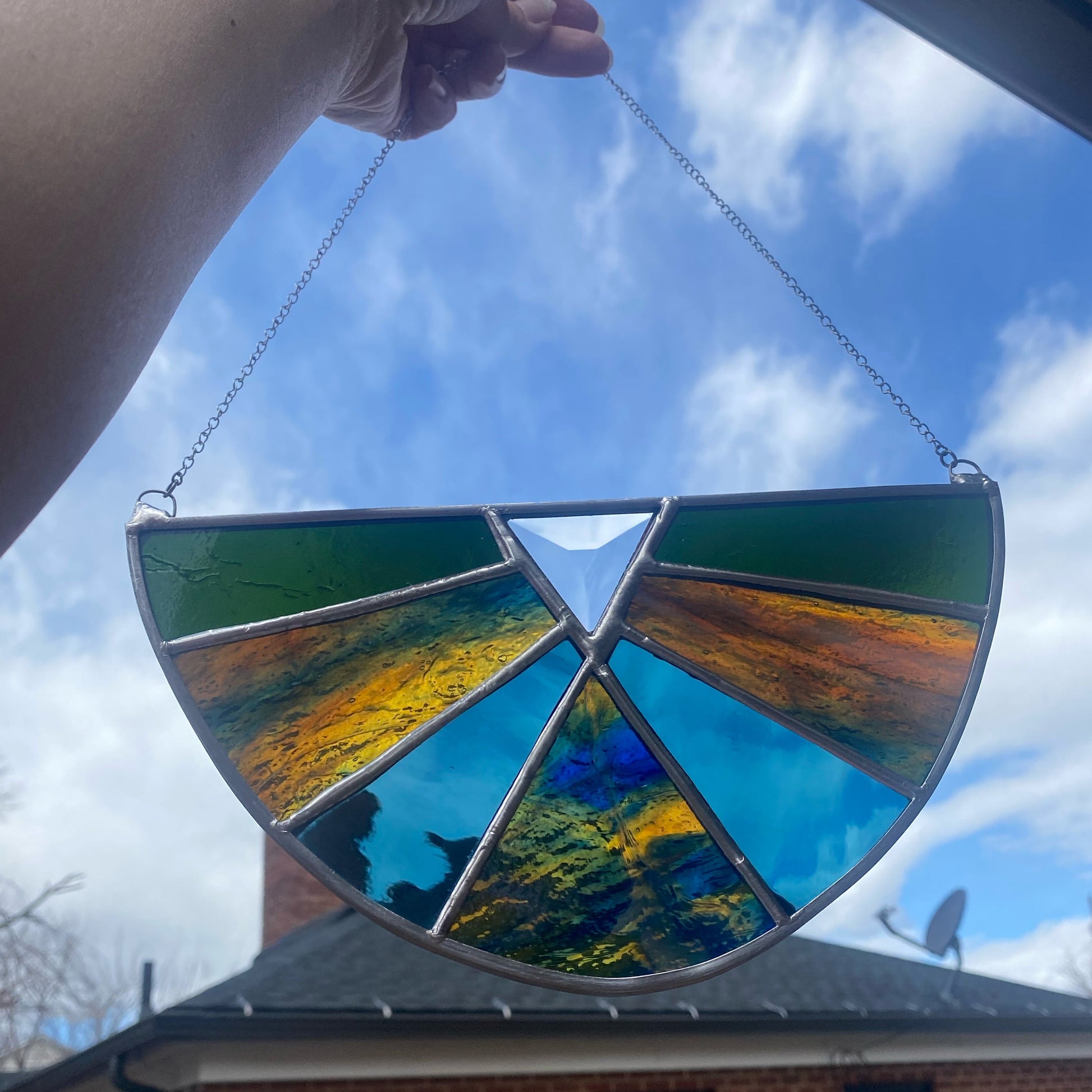A hand holds a semi-circular stained glass window suncatcher outdoors against a blue sky. The suncatcher features multiple colored glass panels in green, blue, yellow, and orange, separated by dark lead lines and suspended by a silver metal chain.