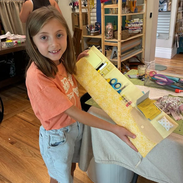 A young girl smiling while holding a long, yellow patterned box in a craft room with shelves of supplies in the background.