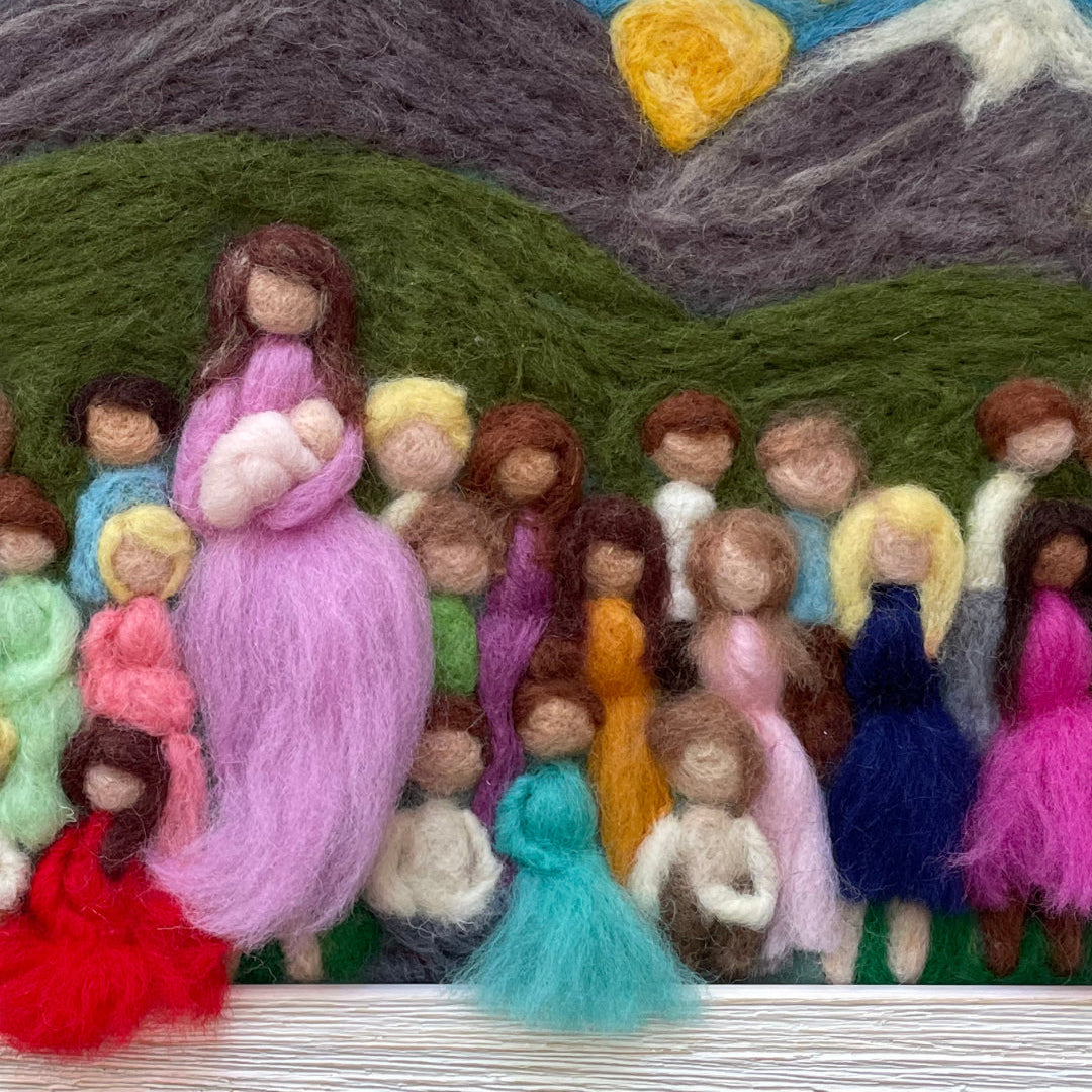 Hand-felted wool art depicting a group of stylized figures in front of mountains and a sun, framed in light wood.
