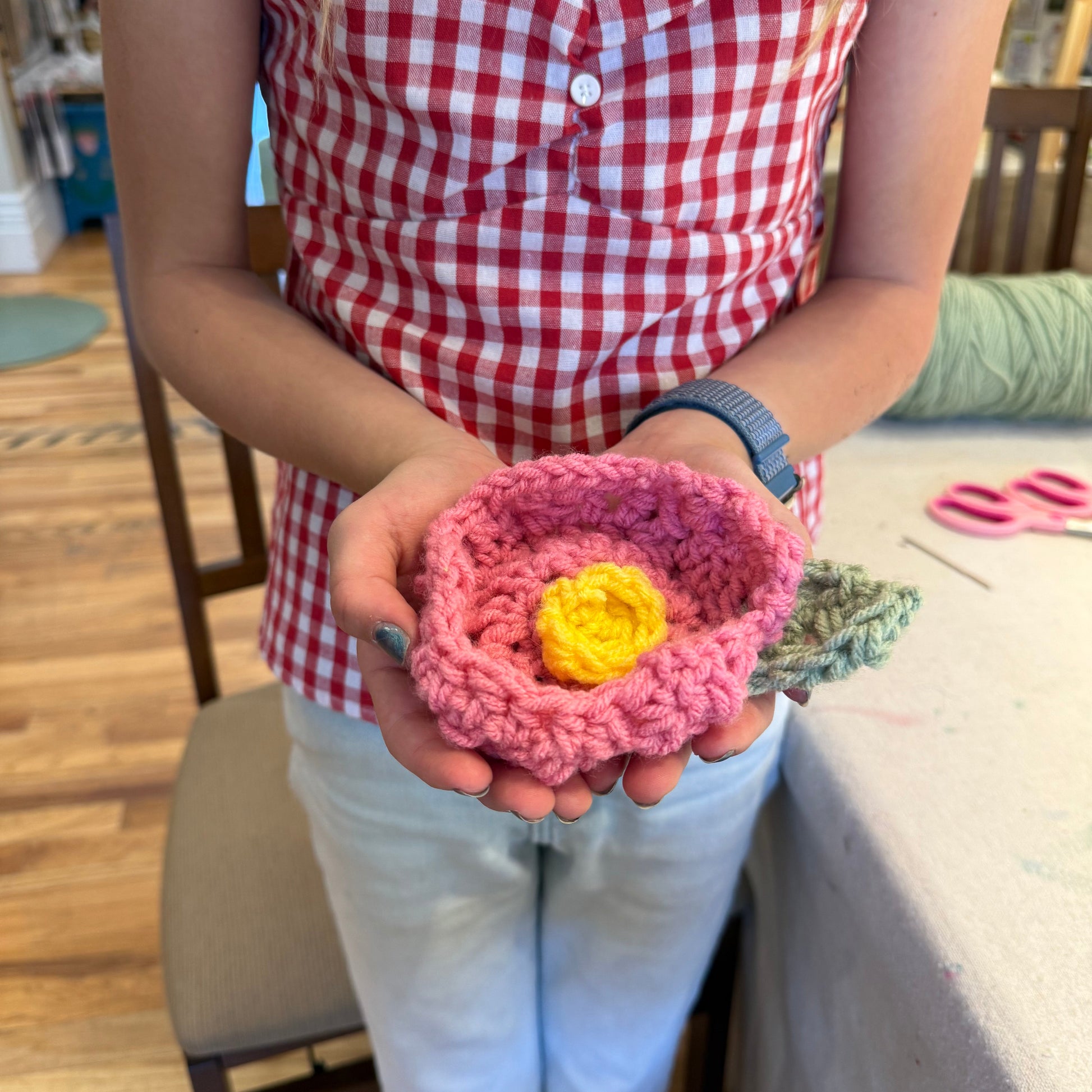 Person holding a pink crocheted flower with a yellow center, wearing a red and white checkered shirt.