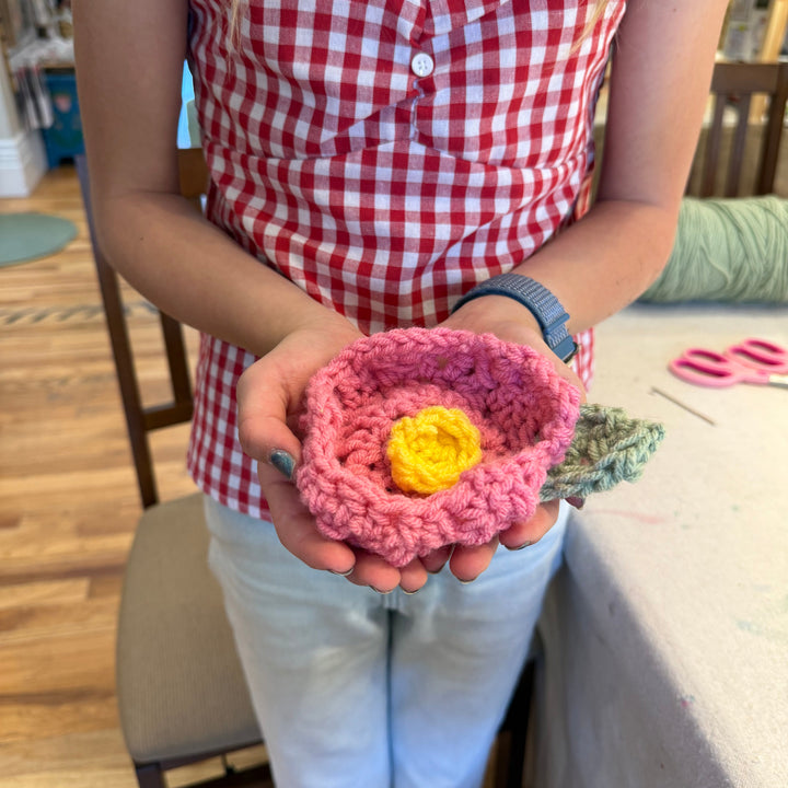 Person holding a pink crocheted flower with a yellow center, wearing a red and white checkered shirt.
