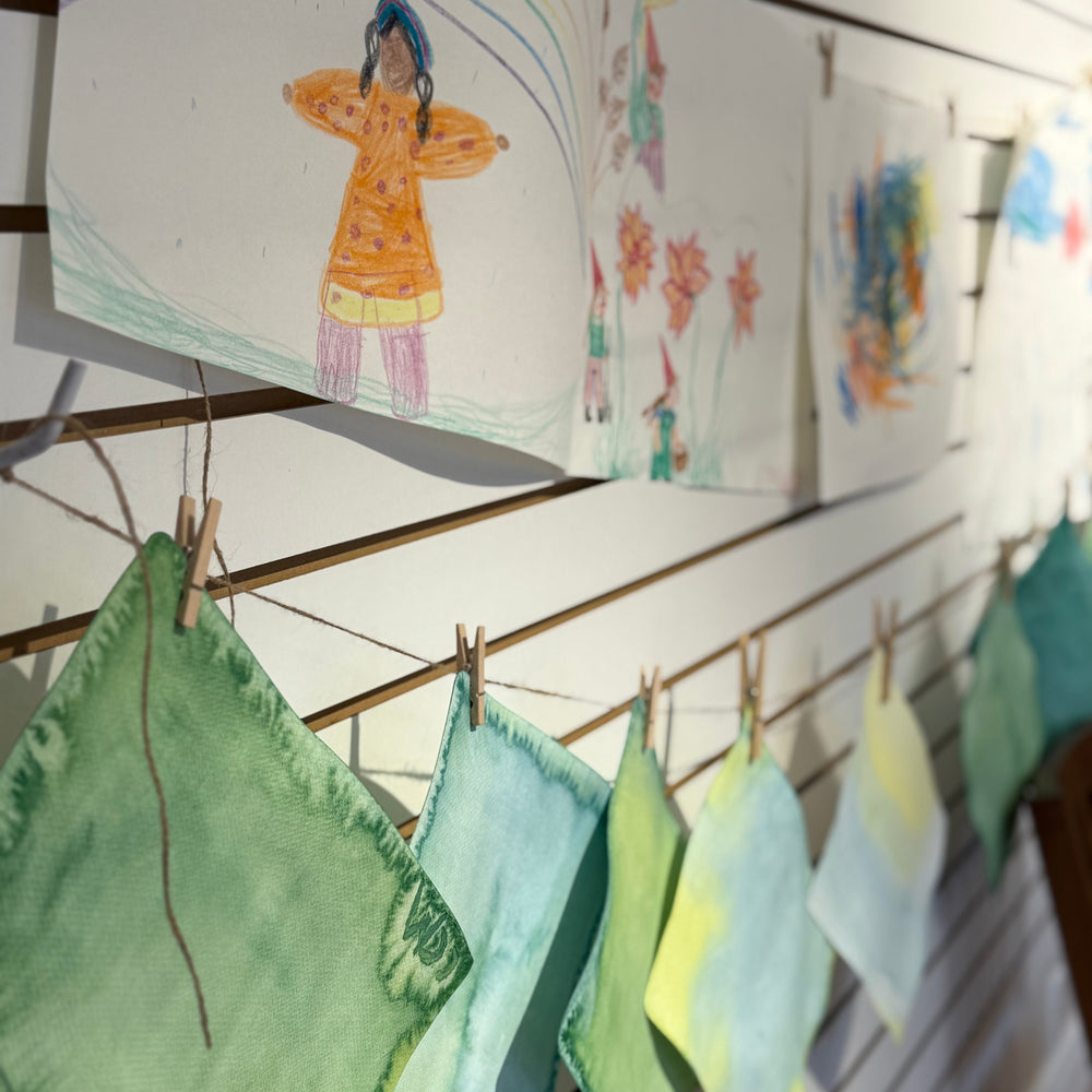 A display of handmade square fabric cloths in various pastel shades of green, blue, yellow, and white, hung on a white slatwall with wooden clothespins. Above the fabrics, children's colored pencil drawings are pinned to the wall.
