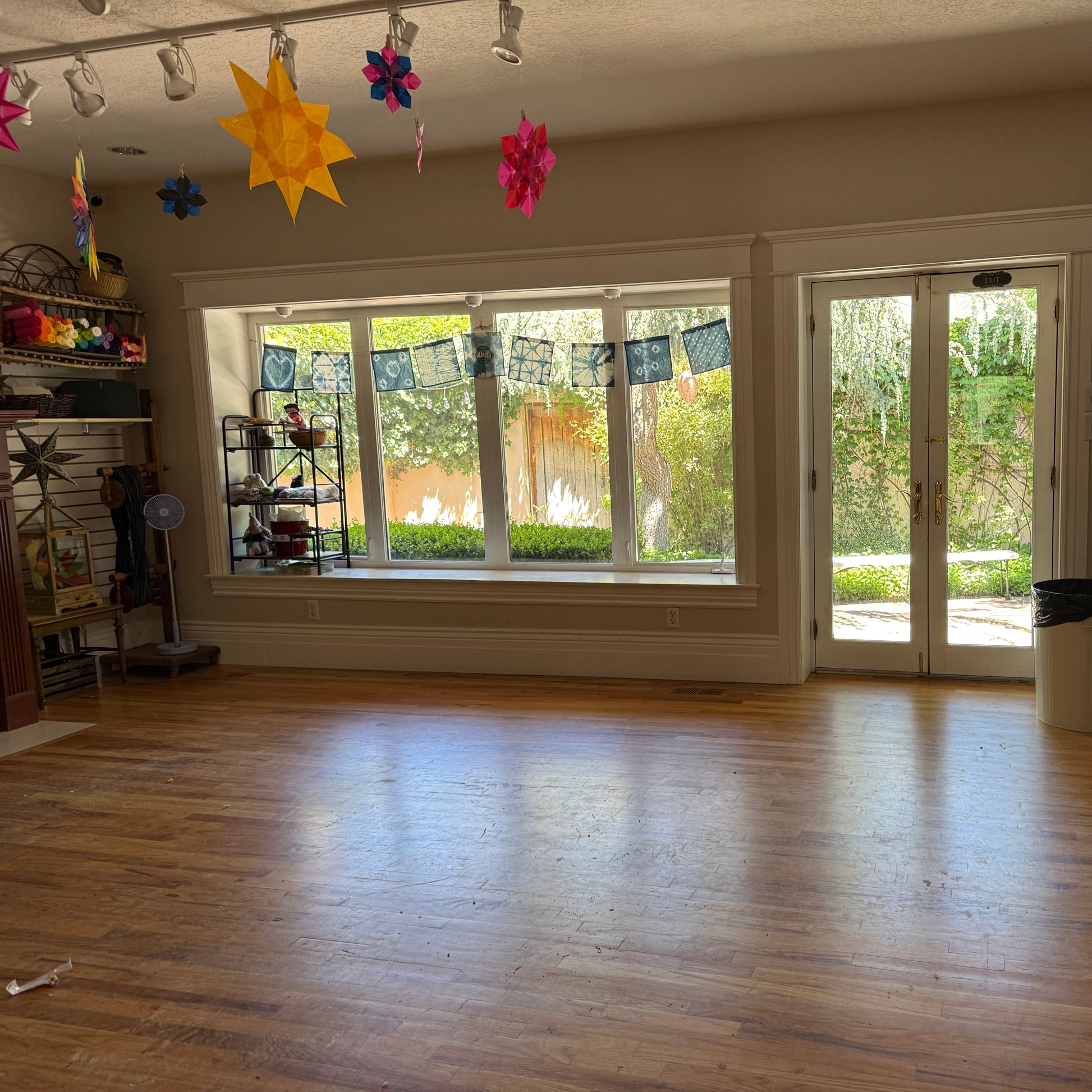 An empty indoor room with hardwood floors, large windows, and glass doors, with colorful hanging star decorations and shelves along the walls.