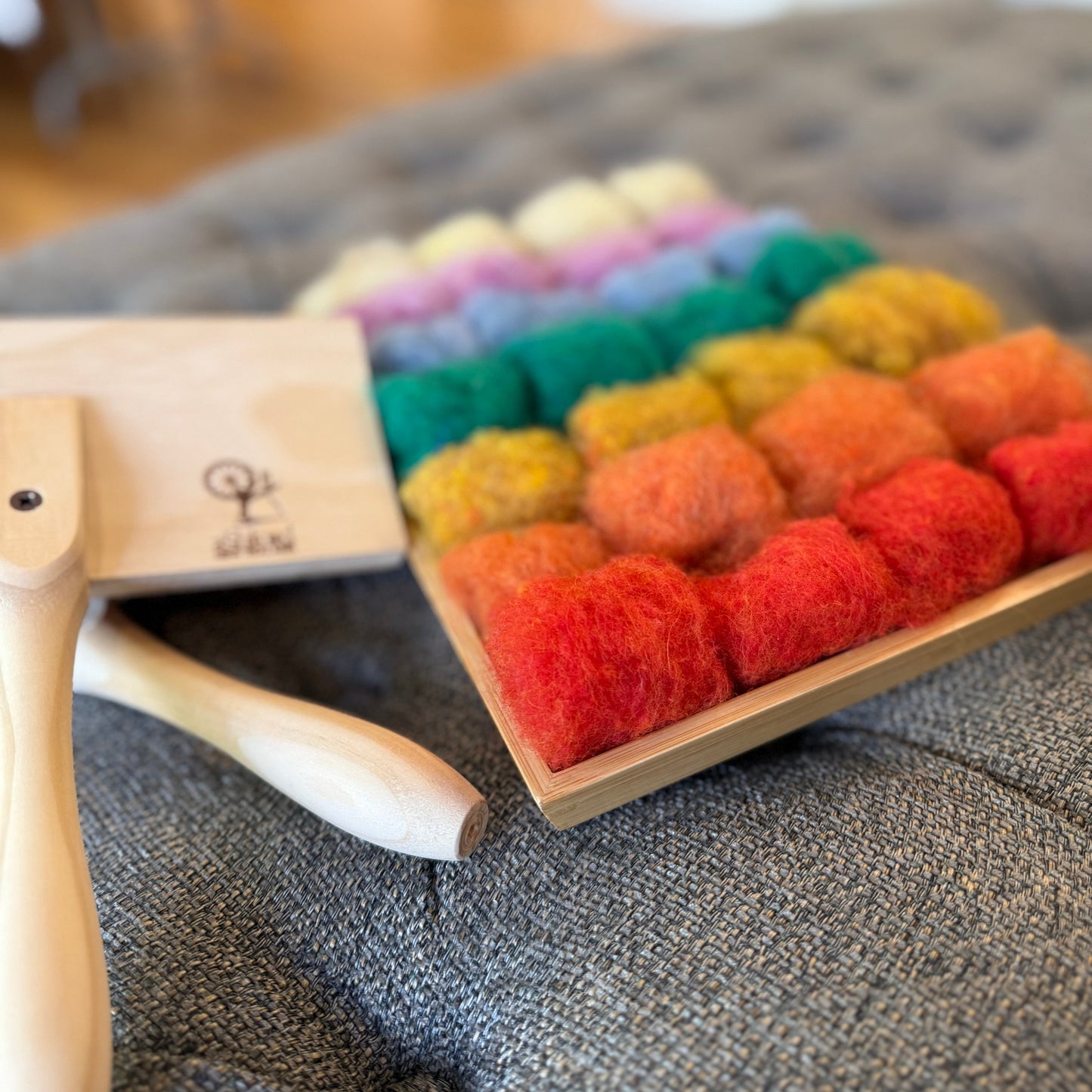 A felted wool craft kit featuring a wooden tray filled with colorful felted wool balls in a rainbow of colors, alongside a wooden tool on a gray tufted bench.