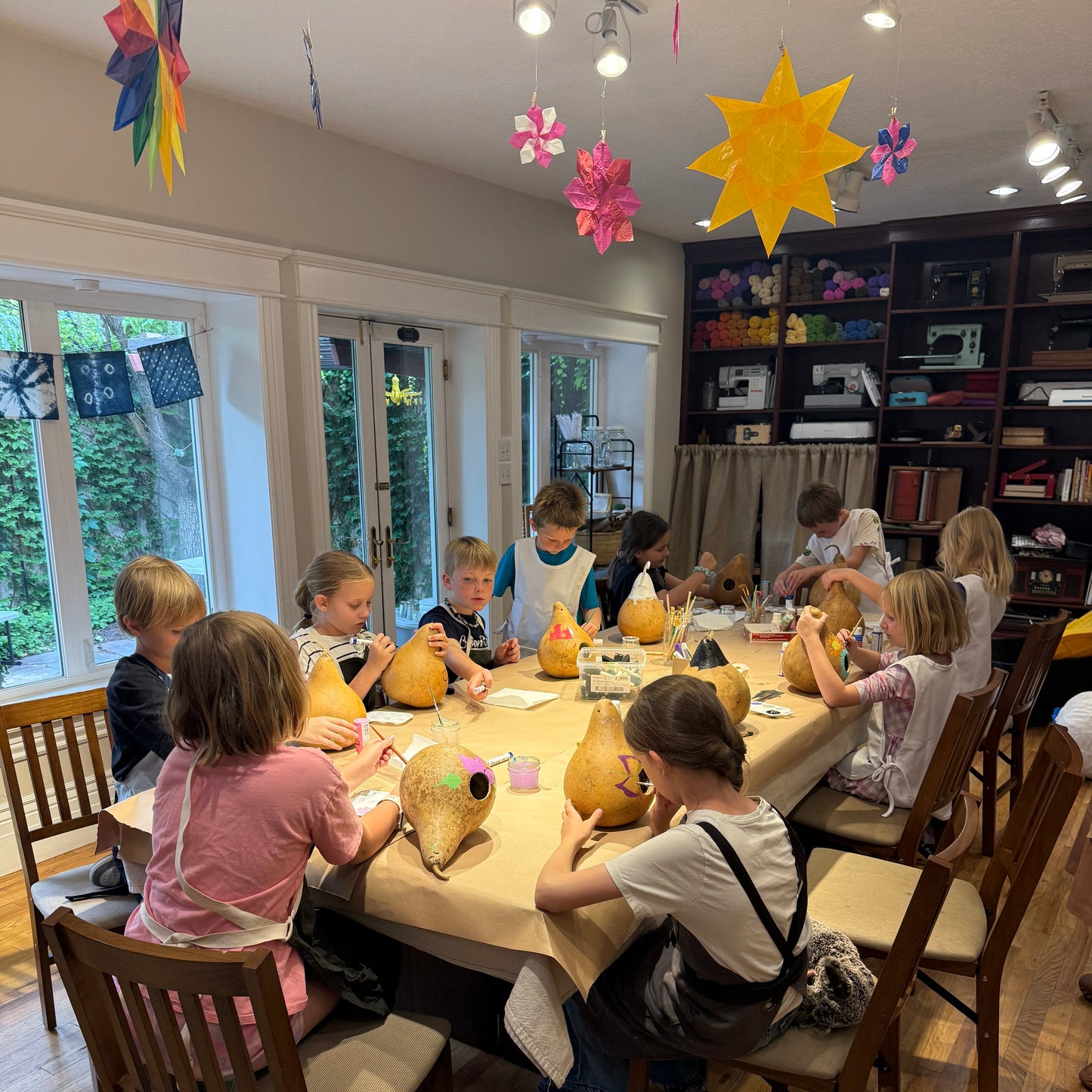 A group of children are gathered around a table in a classroom setting, carving pumpkins. The room features wooden floors, bookshelves, and colorful autumn decorations including a yellow star and paper windmills.