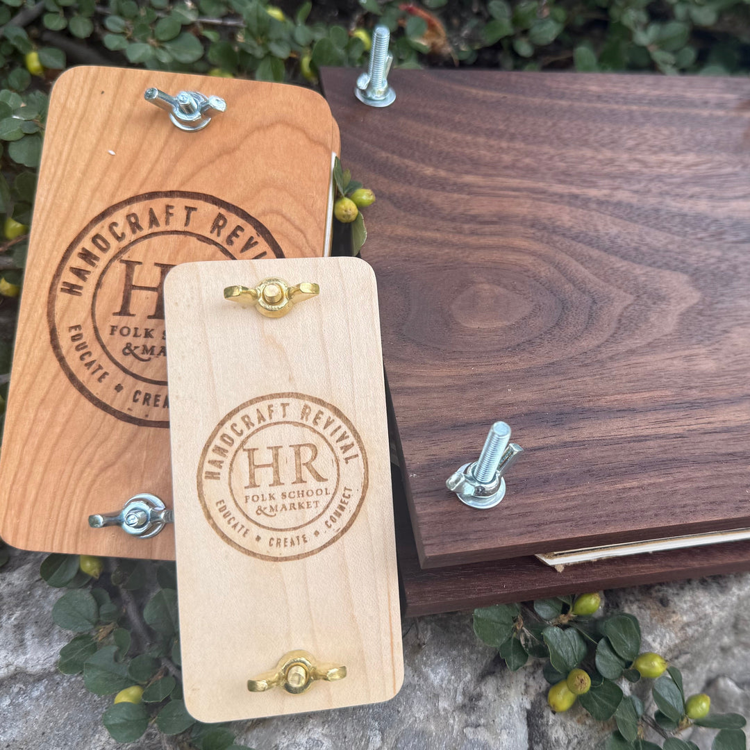 Two wooden keepsake albums or boxes with metal binder-style hardware, one natural wood and one dark walnut wood, displayed outdoors on rocks with green foliage.