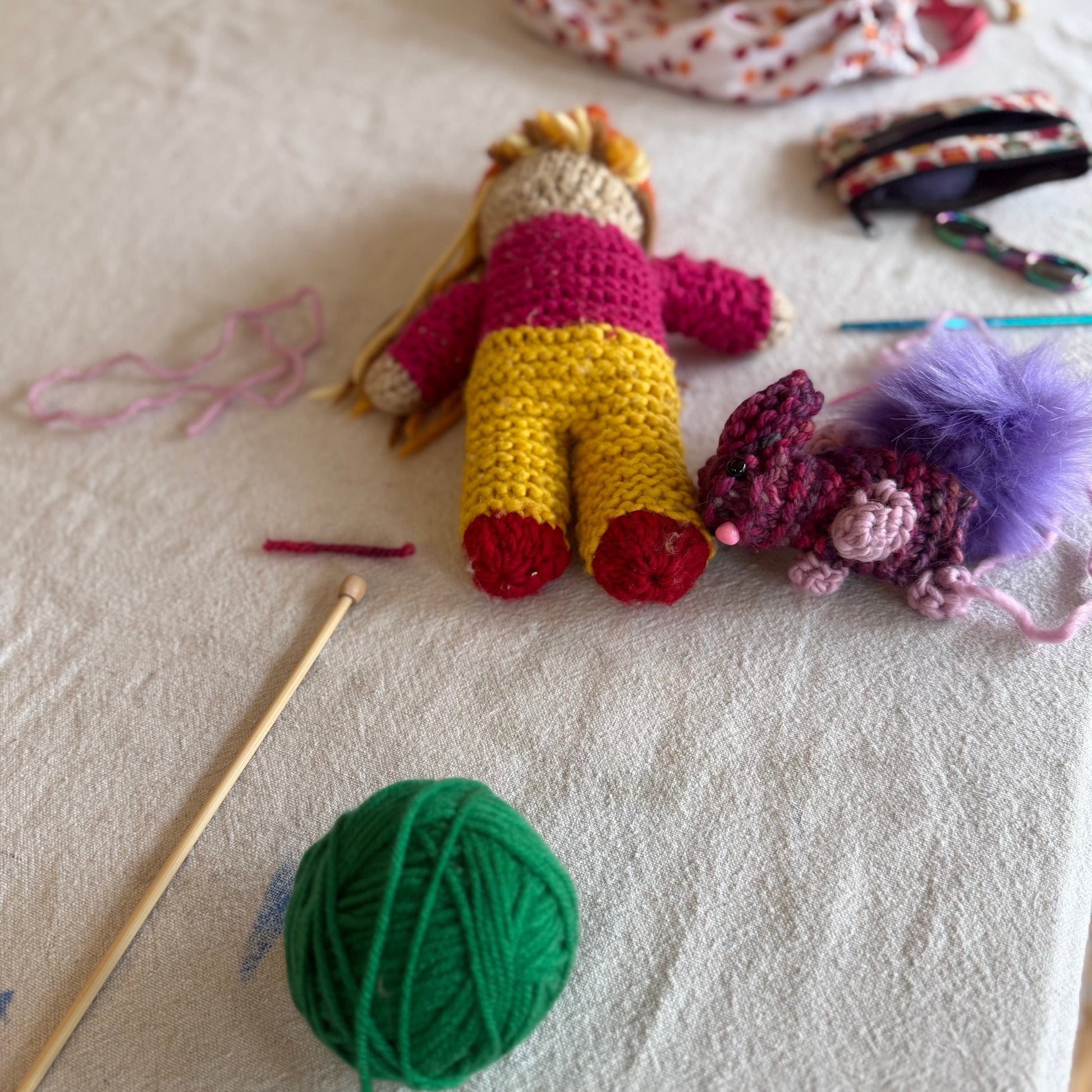 A handmade Knit gnome with yellow hair, a pink top, and yellow pants lies on a white fabric surface, surrounded by knitting tools and yarn including a green ball of yarn, wooden needles, and other colorful crochet items.