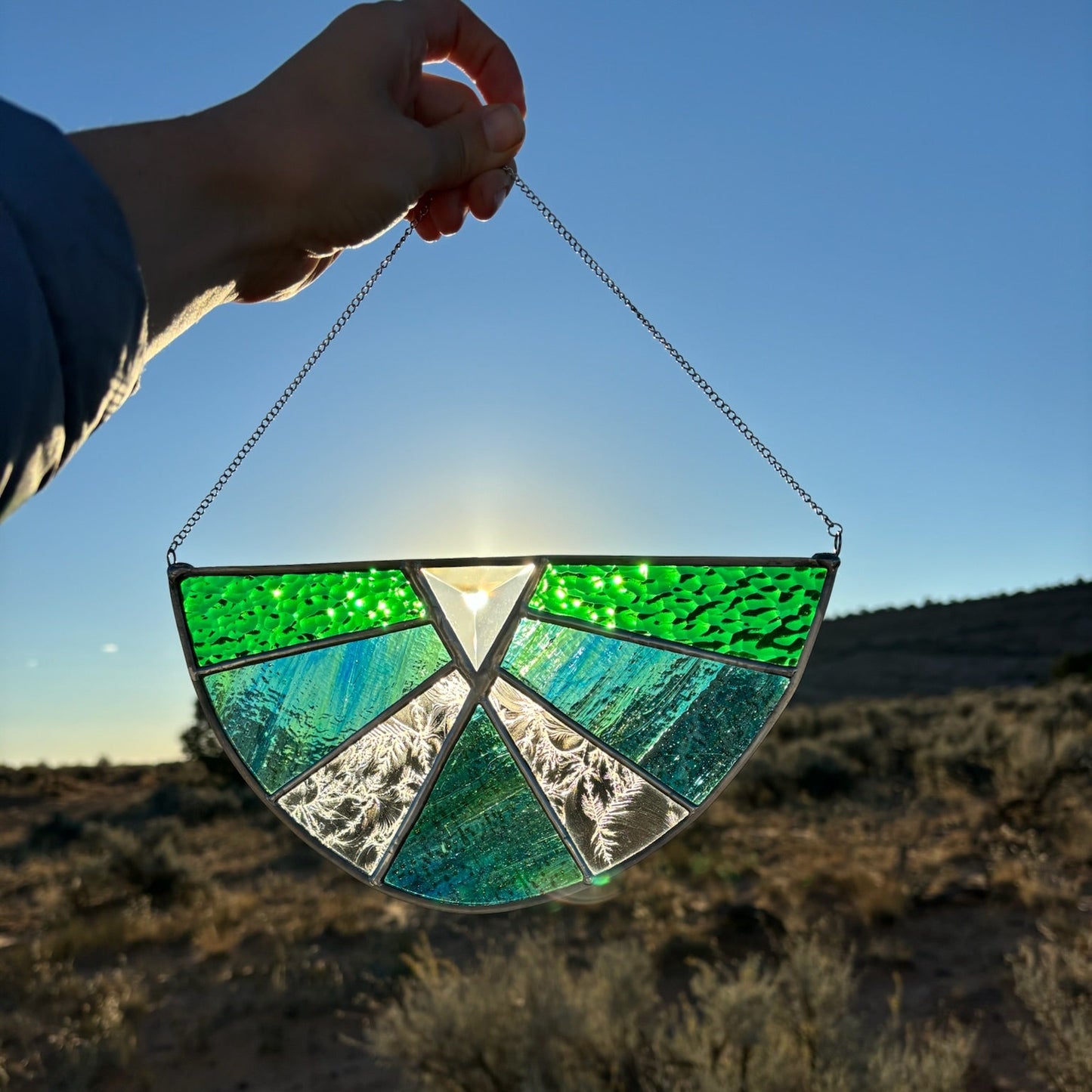 A hand holds a semicircular stained glass suncatcher outdoors in a desert landscape at sunset. The suncatcher features textured bright green and teal-blue glass panels with silver metal separators and a hanging chain.