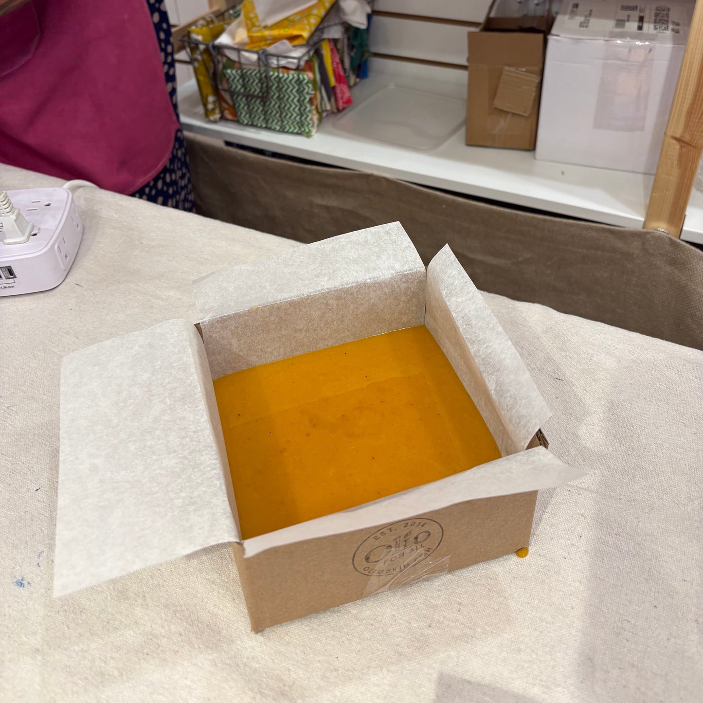 An open kraft cardboard box lined with white tissue paper, filled with a bright orange-yellow liquid, set on a white tablecloth in a retail or workshop environment.