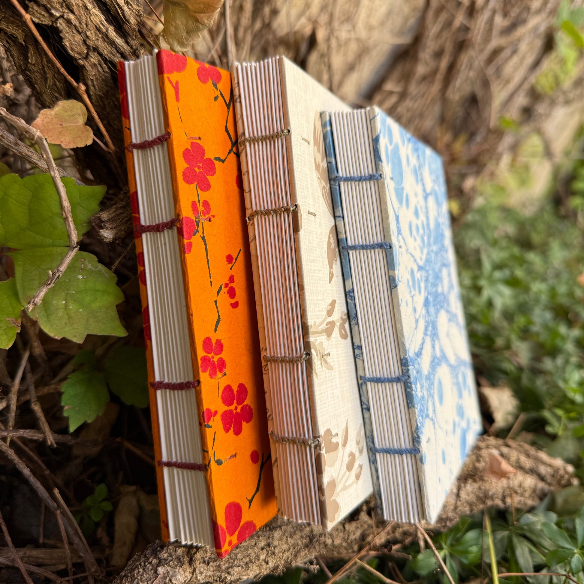 Three handmade bound books with floral and patterned covers are propped against a tree trunk outdoors, surrounded by green leaves and natural branches.