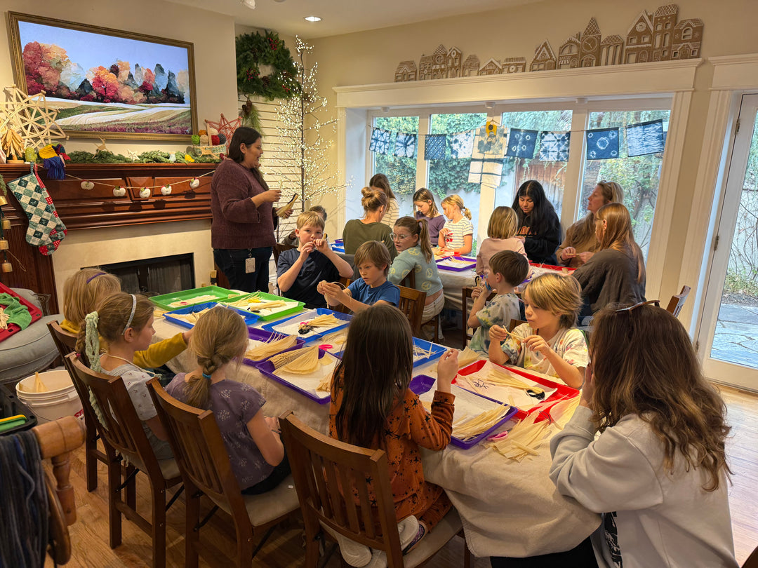 A group of children and adults gathered around a table in a decorated indoor room, working on craft or activity projects with papers, scissors, and colorful materials. A woman stands at the head of the table speaking to the group, with windows, holiday decorations, and wall art visible in the background.