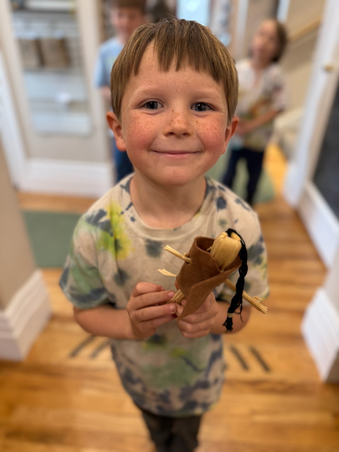 A young boy with freckles stands in a hallway holding a small brown felt toy pouch with a black ribbon.