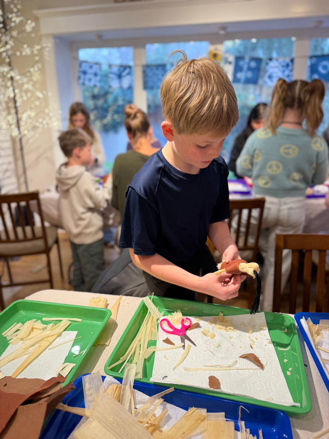 A young boy in a navy blue t-shirt is actively participating in a craft activity at a table, holding a piece of wood while trays of wooden shavings, scissors, and paper pieces are spread out in a brightly lit indoor room.
