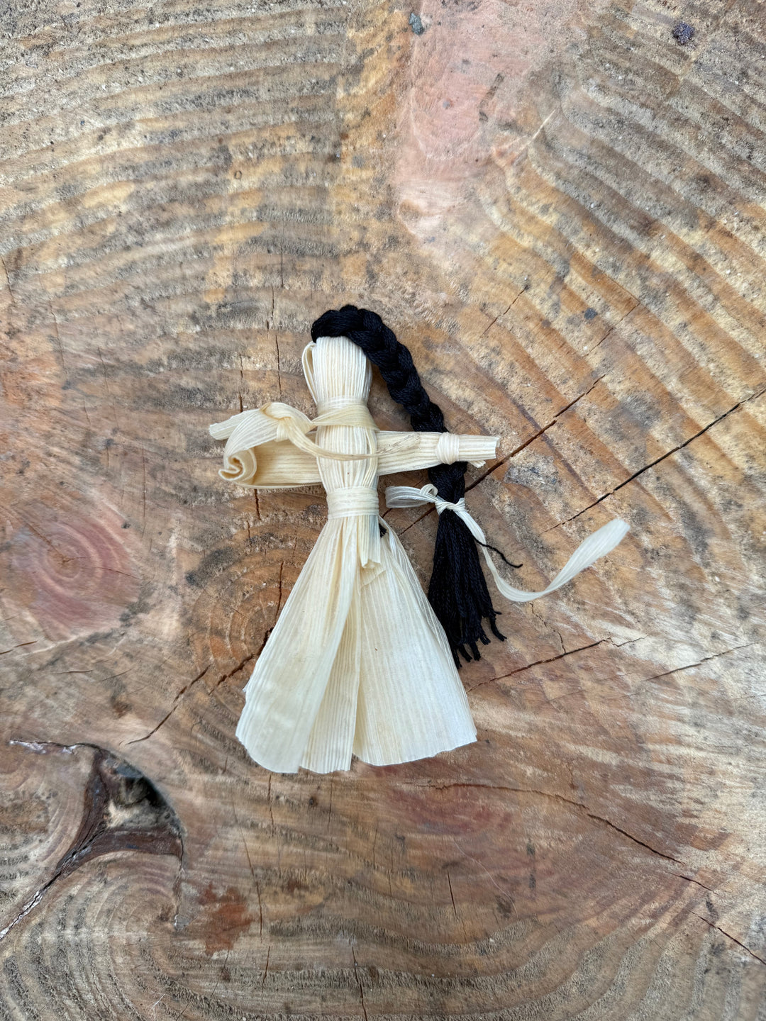 A small handmade doll made from natural materials, featuring a pale beige body crafted from dried corn husks and a long black braided hair-like element, placed on a textured wood surface.