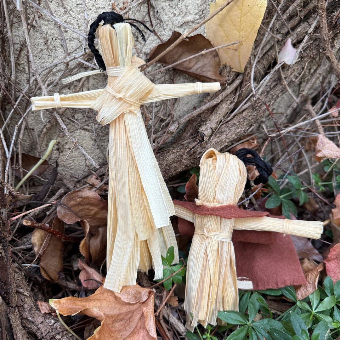 Two small handmade reed or straw dolls with black yarn hair are placed outdoors among dry leaves, twigs, and green plants against a textured wall.