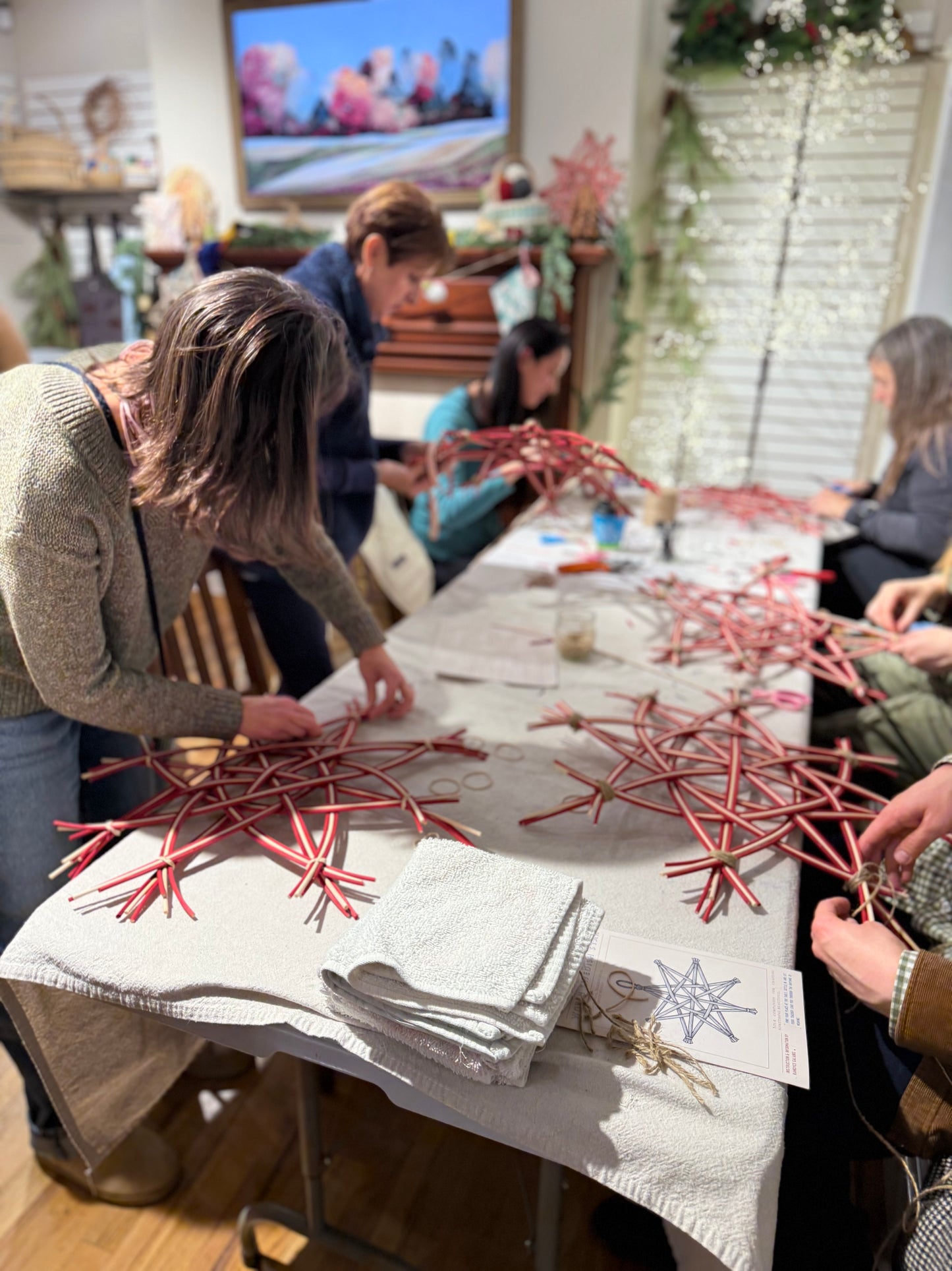 Several people gathered around a table indoors, assembling red metal star-shaped ornaments or decorations on a white cloth-covered table. Papers and tools are visible on the table, and a decorated room with shelves and wall art appears in the background.
