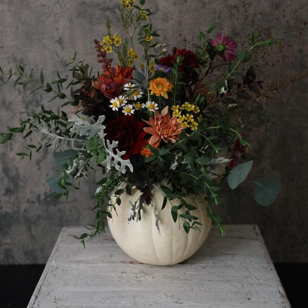 An autumn floral arrangement in a ribbed cream-colored pumpkin vase, featuring a mix of red, orange, yellow, and white wildflowers with green and silver foliage, displayed on a distressed white wooden table against a textured gray wall.