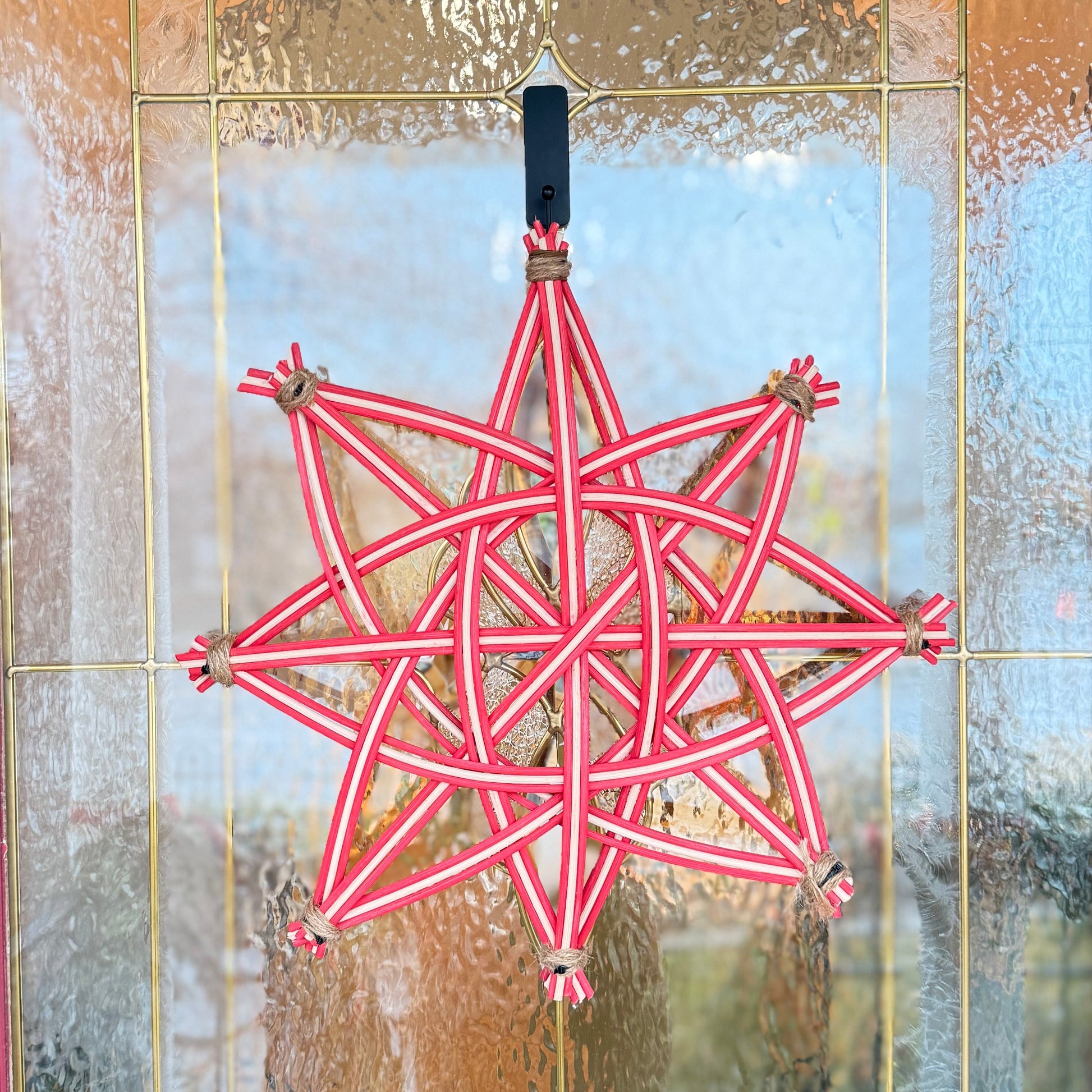 A red and white braided star-shaped hanging ornament with small bells at the points, displayed on a textured glass front door.