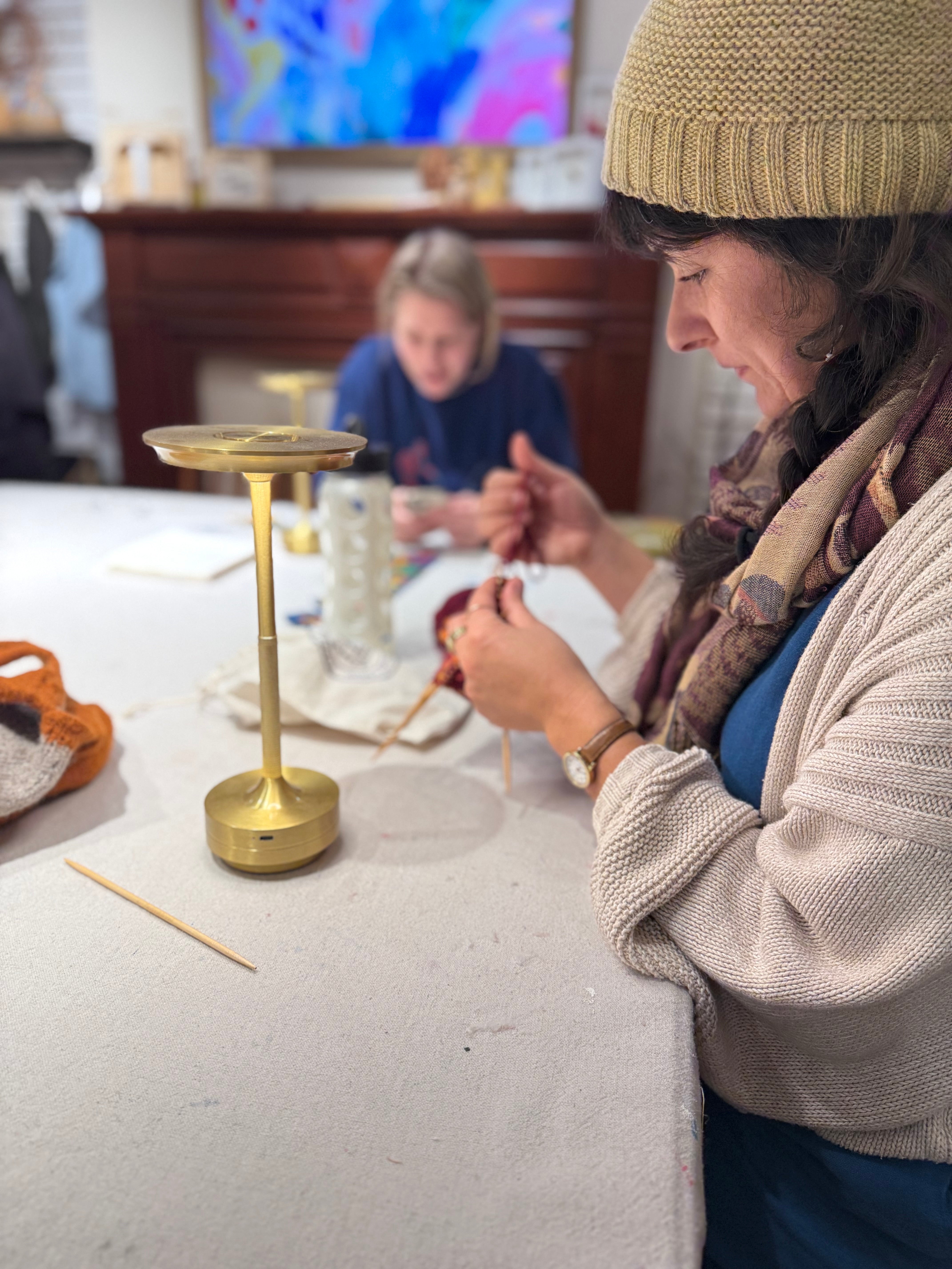 A woman in a beige knit beanie and cream cardigan is shown in a cozy indoor setting, focused on crocheting or knitting at a table with a brass candle holder. Another person is visible in the background.