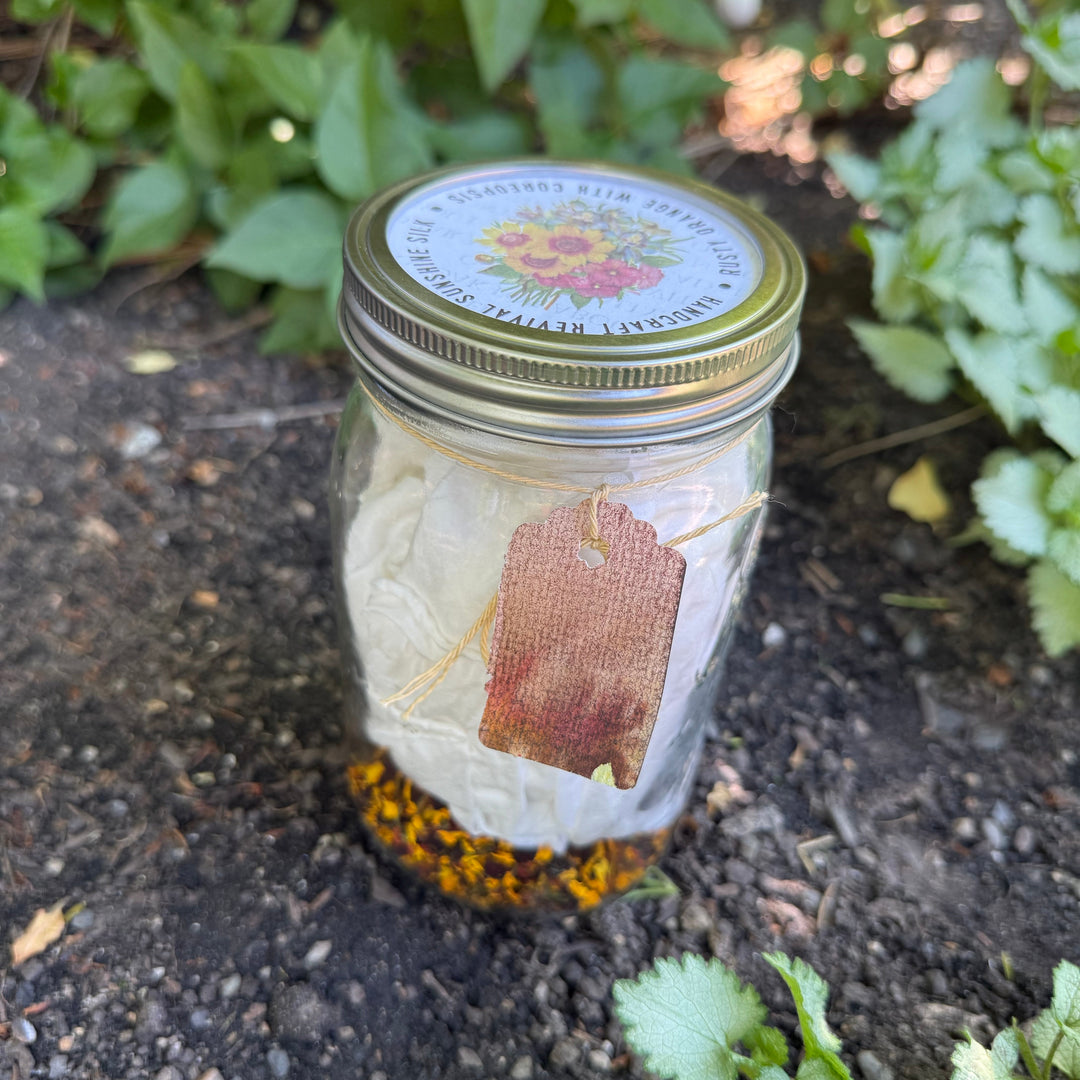 A clear glass mason jar with a gold metal lid sits on dark garden soil surrounded by green plants. The jar contains white folded paper towels or tissue paper, a small burlap tag tied with string, and a layer of dried orange flowers at the bottom.