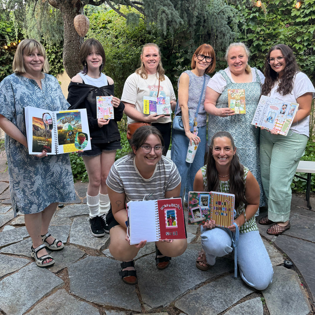 Seven women posing together outdoors on a stone patio, each holding colorful printed cards or small books. Trees, hanging string lights, and greenery are visible in the background.