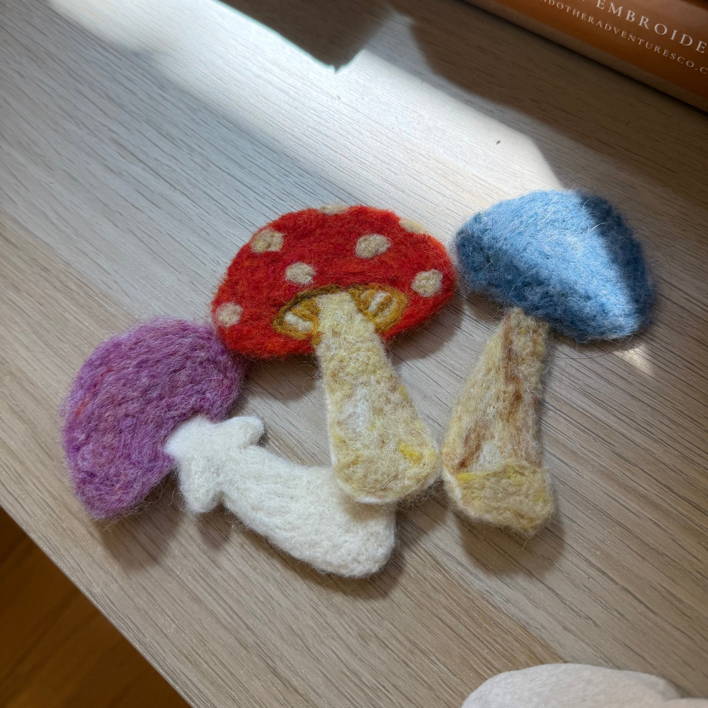Four hand-felted mushroom-shaped items in red, blue, purple, and white with beige stems, arranged on a light wood surface with boxes in the background.