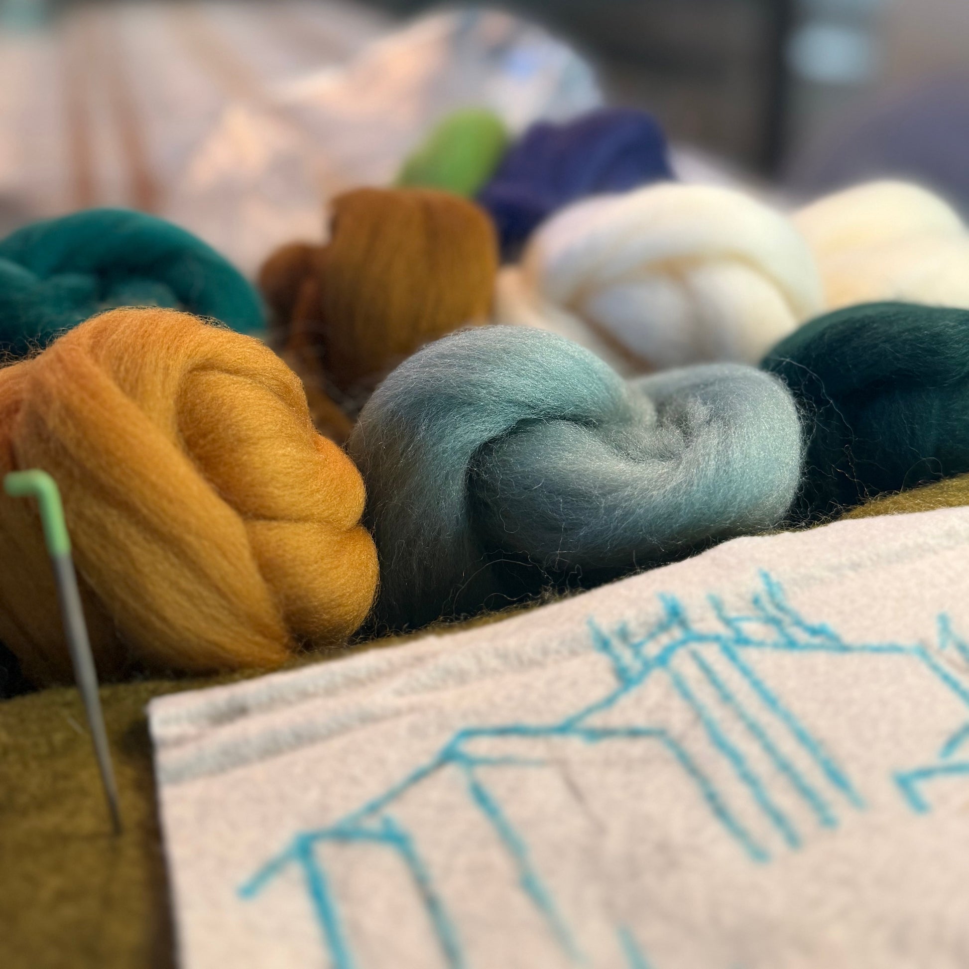 A close-up of colorful wool fibers and yarn skeins arranged on a table, with a white fabric sample in the foreground showing blue stitched lines. The background is softly blurred, suggesting a crafting or studio environment.
