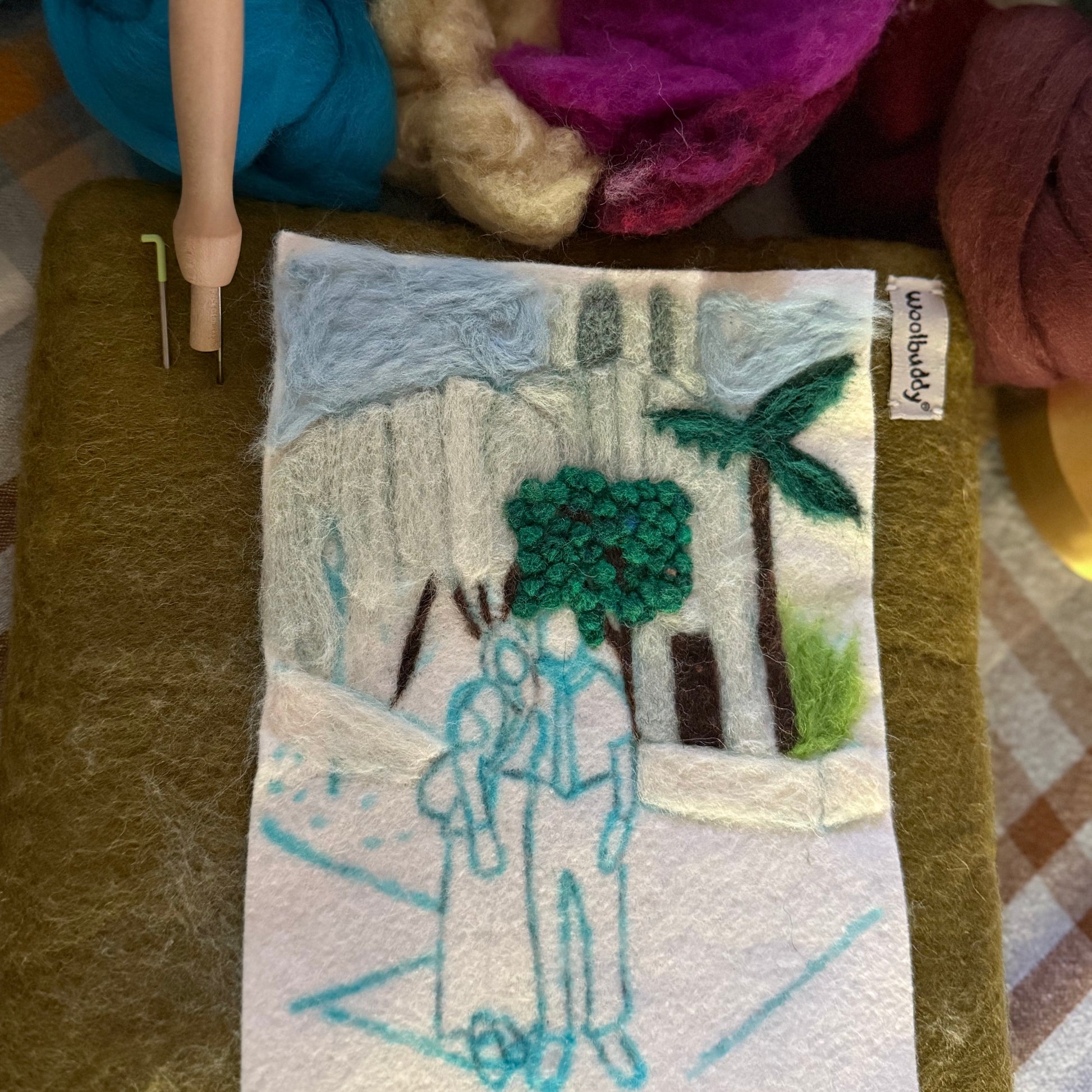 A handmade felt art piece depicting a couple under a palm tree, displayed on a felt pad with wool roving and a needle loom in the background.