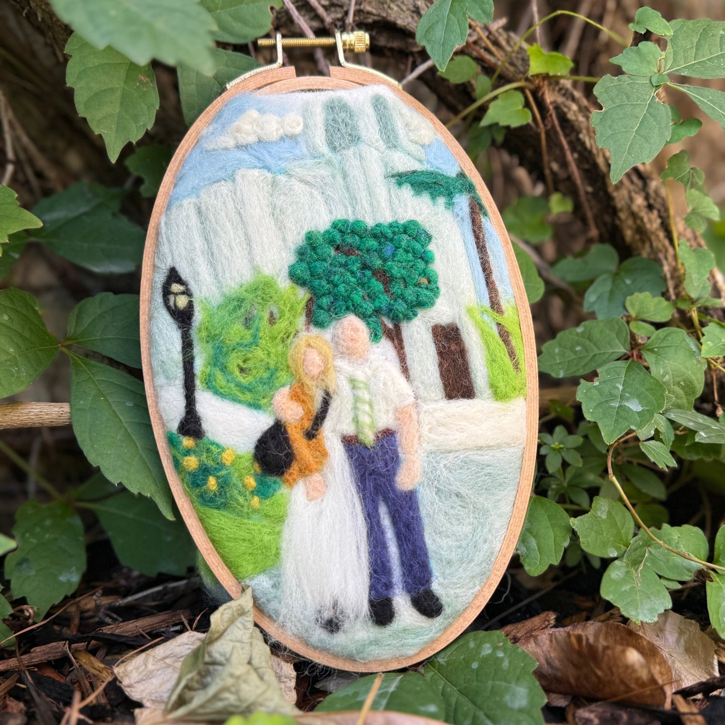 An oval felt needlework hanging ornament depicting a couple in a park, suspended by a string from a tree branch outdoors among green leaves and brown leaves on the ground.