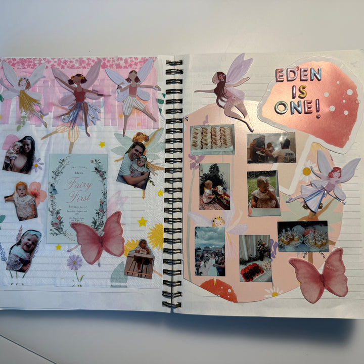 An open spiral-bound journal spread showing a decorated fairy-themed collage with pastel pink and peach sections, butterfly stickers, small photo inserts, stars, and the text &