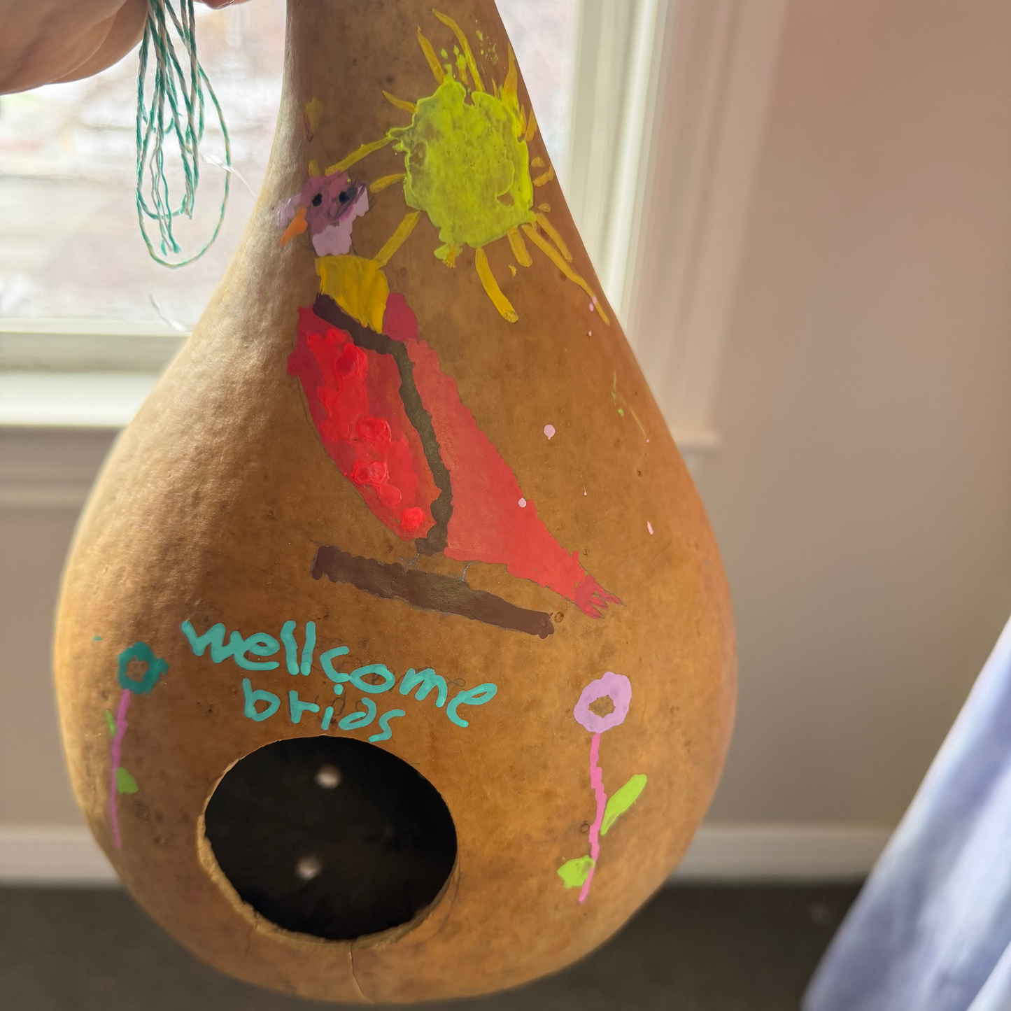 A hand holds a decorated natural gourd with a birdhouse opening. The gourd is painted with a red bird, a yellow sun, flowers, and the handwritten text 'wellcome brian'.