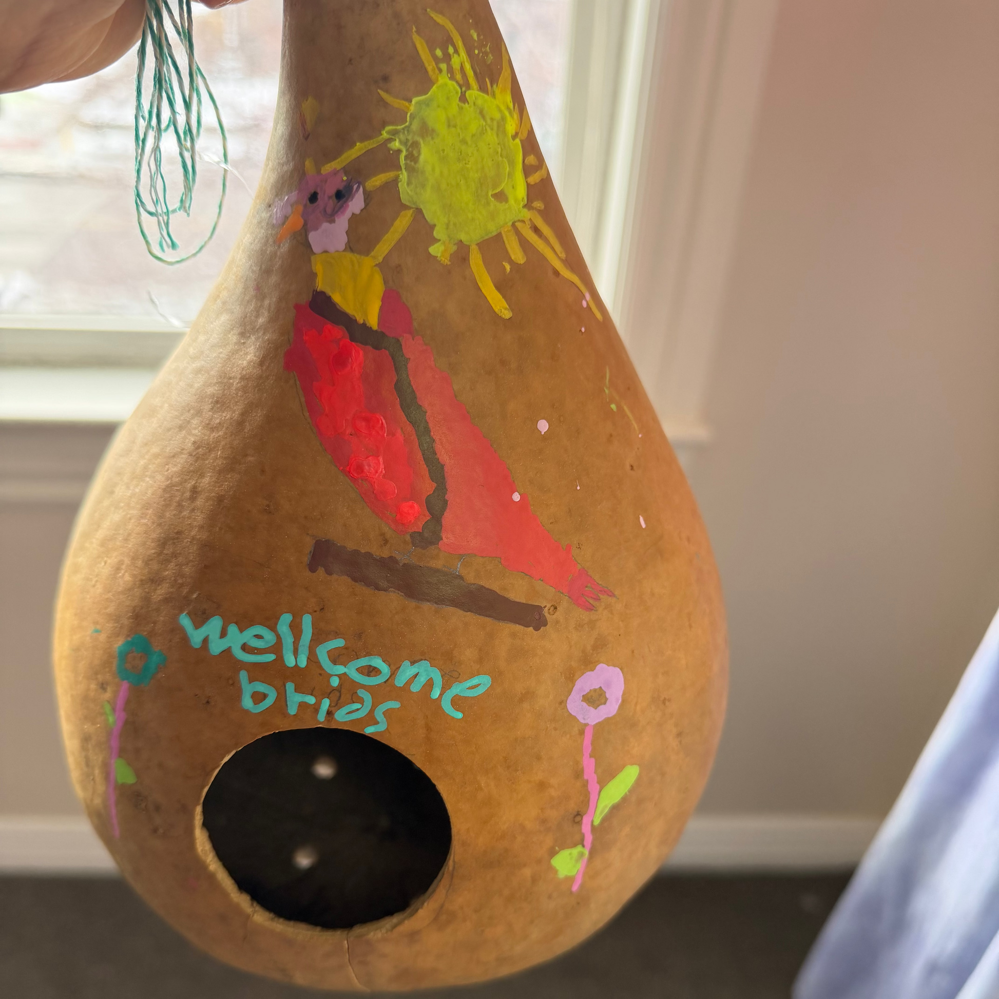 A hand holds a decorated natural gourd with a birdhouse opening. The gourd is painted with a red bird, a yellow sun, flowers, and the handwritten text 'wellcome brian'.
