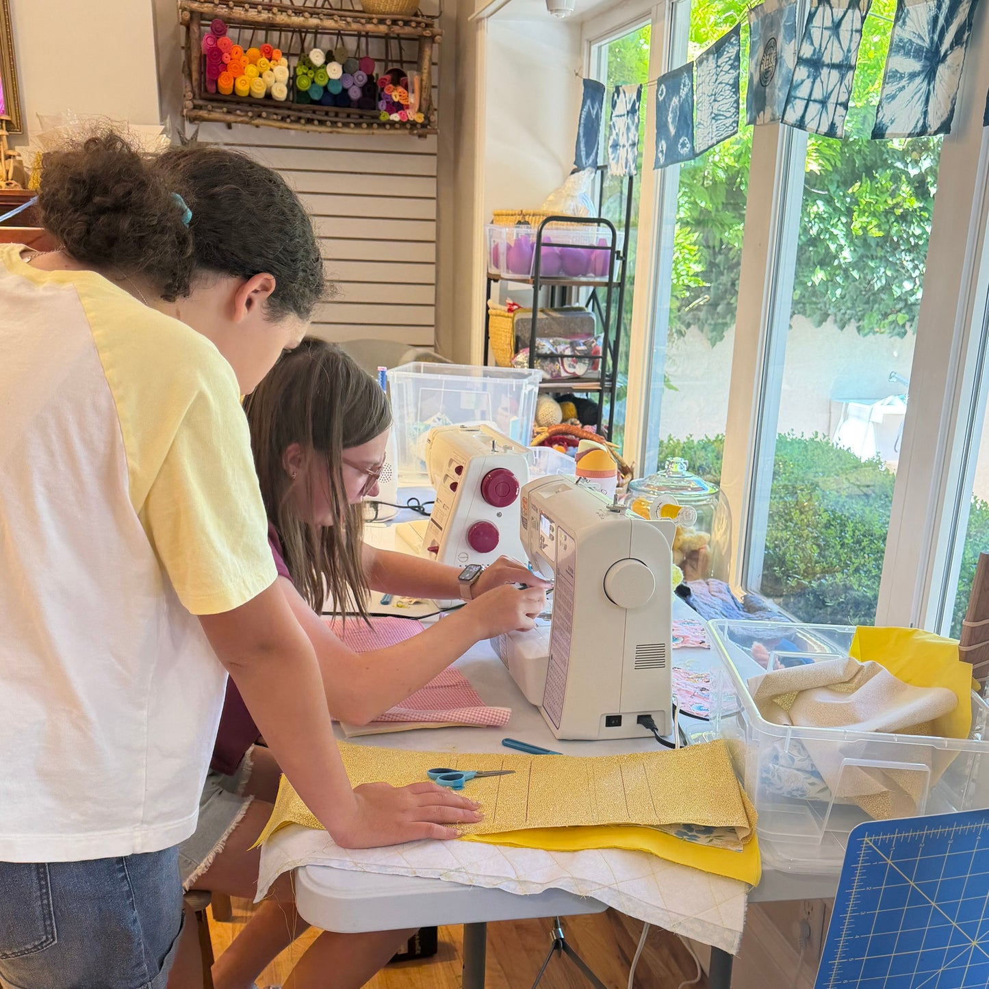 Two girls in a bright craft room using a white sewing machine, with one girl guiding the fabric and the other observing closely.