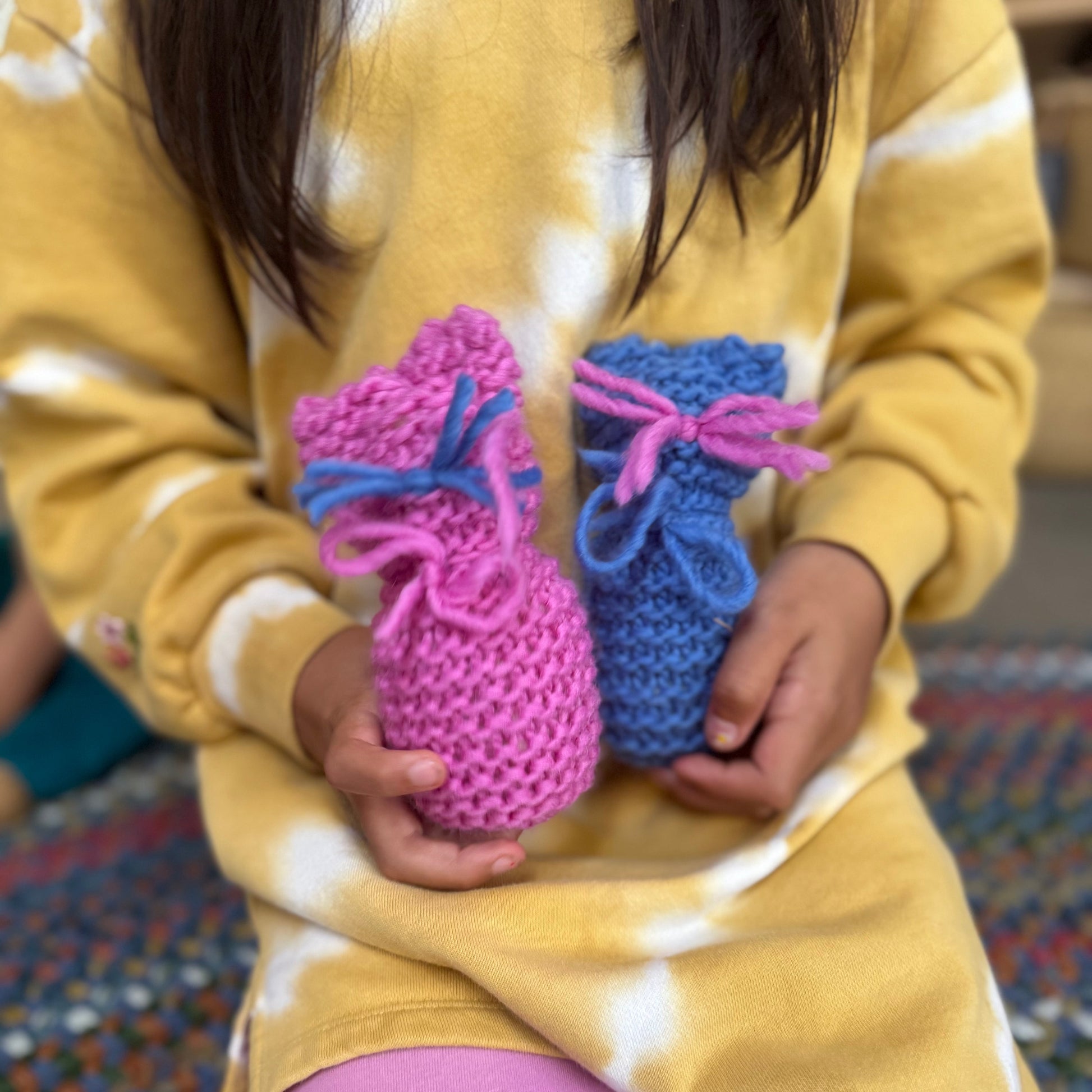 Child holding two knitted booties, one pink and one blue, in a yellow and white patterned shirt.