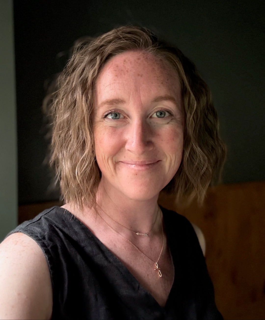 A close-up portrait of a smiling woman with shoulder-length wavy light brown hair, wearing a black sleeveless top and delicate gold necklaces, set against a dark background.