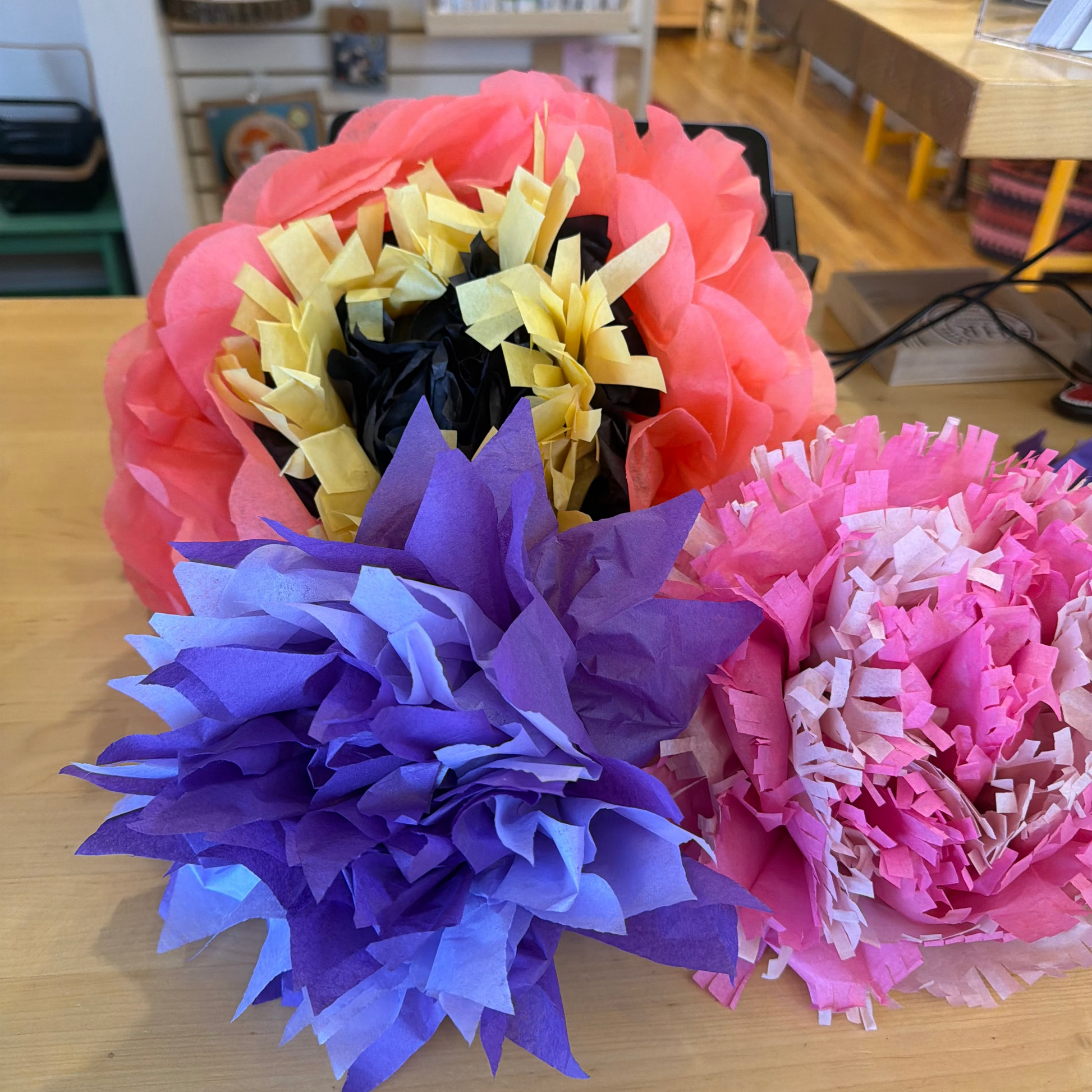 Three decorative tissue paper flower pom-poms in coral pink, purple, and pink, arranged on a light wood tabletop in a store setting.