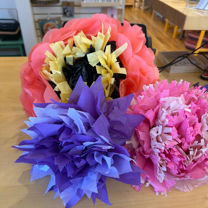 Three decorative tissue paper flower pom-poms in coral pink, purple, and pink, arranged on a light wood tabletop in a store setting.