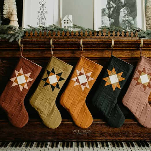 Five quilted Christmas stockings in earthy brown, olive, mustard, black, and dusty pink hang from a wooden mantel decorated with greenery, framed art, and a small white house, with a piano keyboard visible at the bottom.