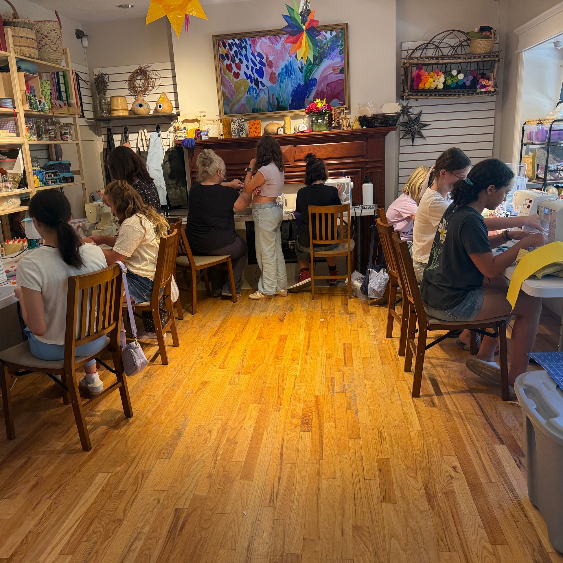 A bright, wood-floored craft studio with several people seated at sewing machines, shelves of supplies along the walls, and a colorful framed artwork hanging above a wooden cabinet.