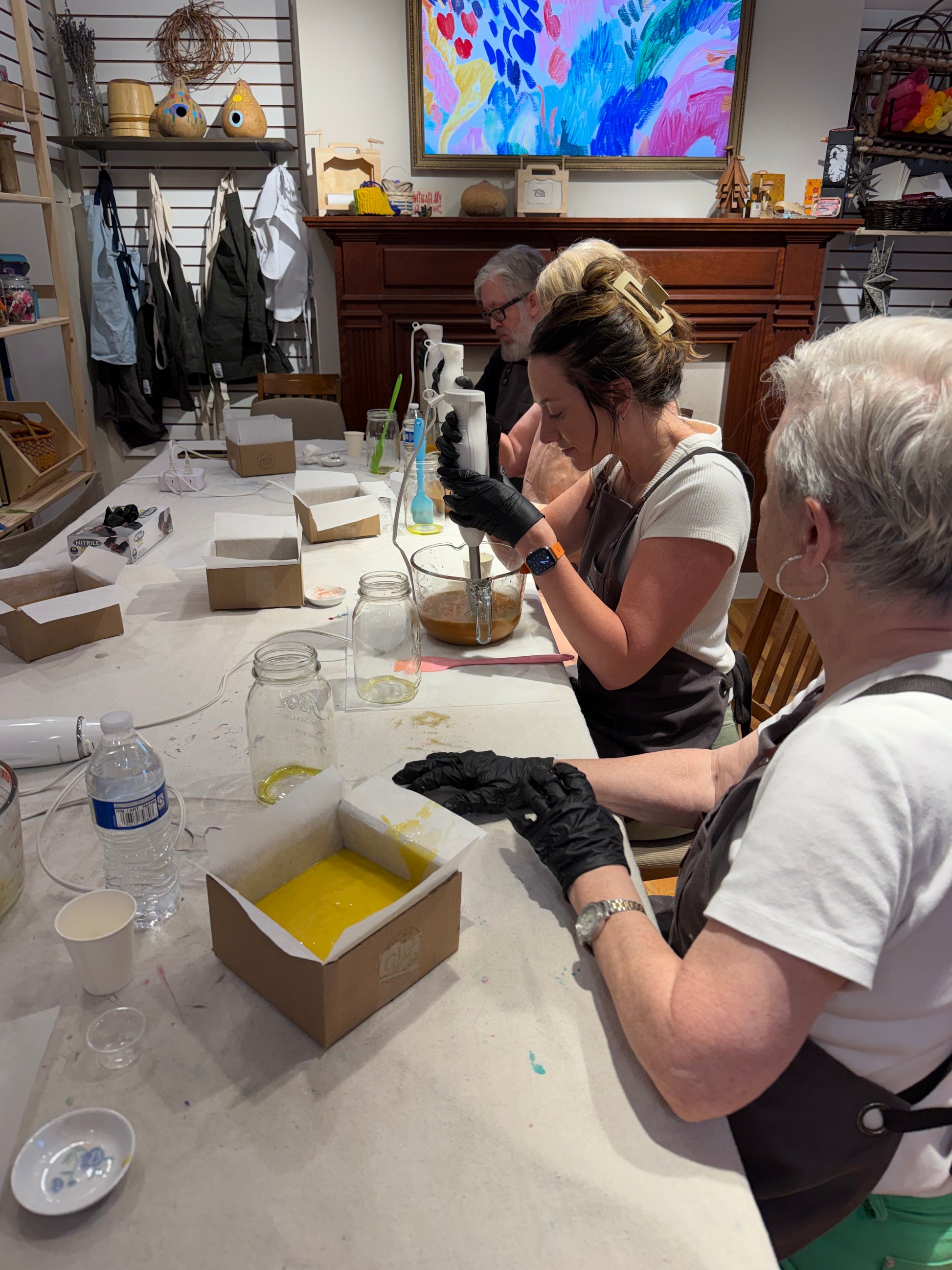 Three people wearing aprons and black gloves sit at a long table in a craft studio, working with glass jars, mixing tools, and a yellow liquid in cardboard boxes. A colorful abstract painting hangs on the wall in the background.