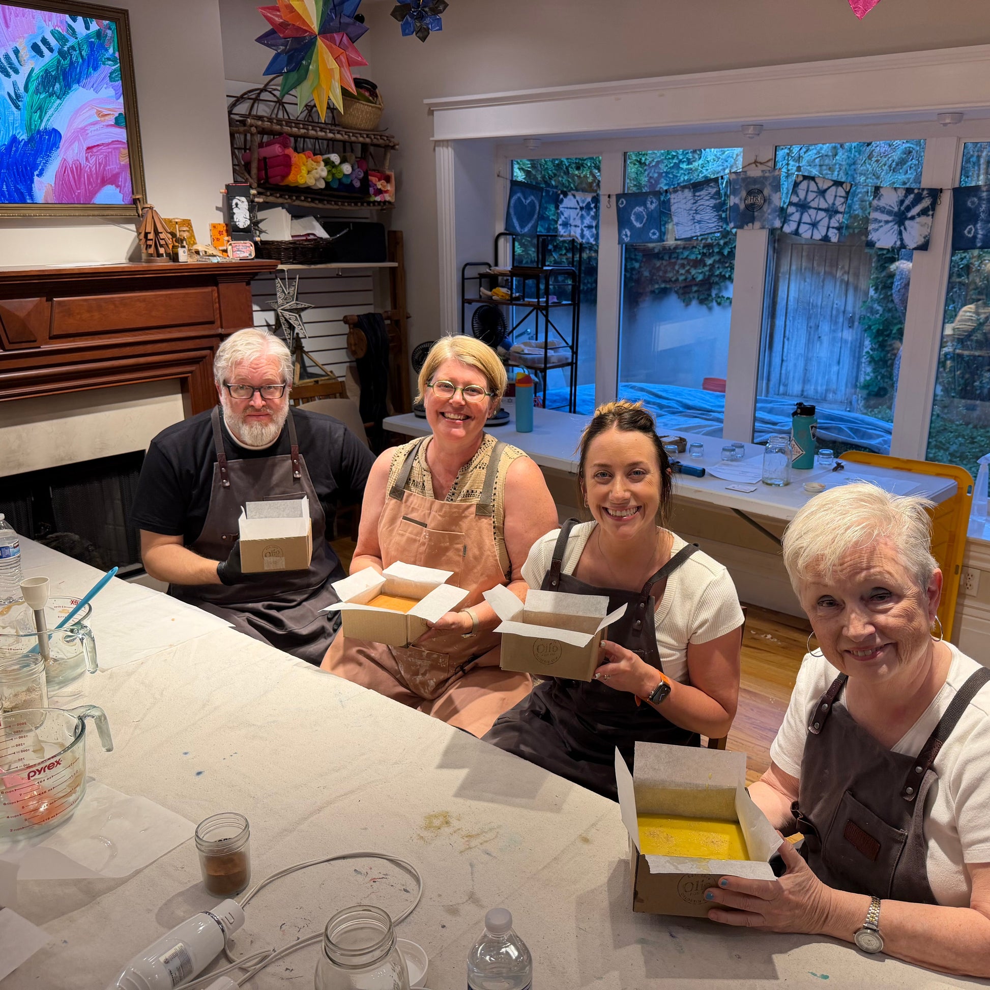 Four people wearing aprons sit around a table in a home studio, holding small boxes containing colorful painted tiles. The room has a fireplace, a large colorful painting on the wall, and windows looking out onto a garden.