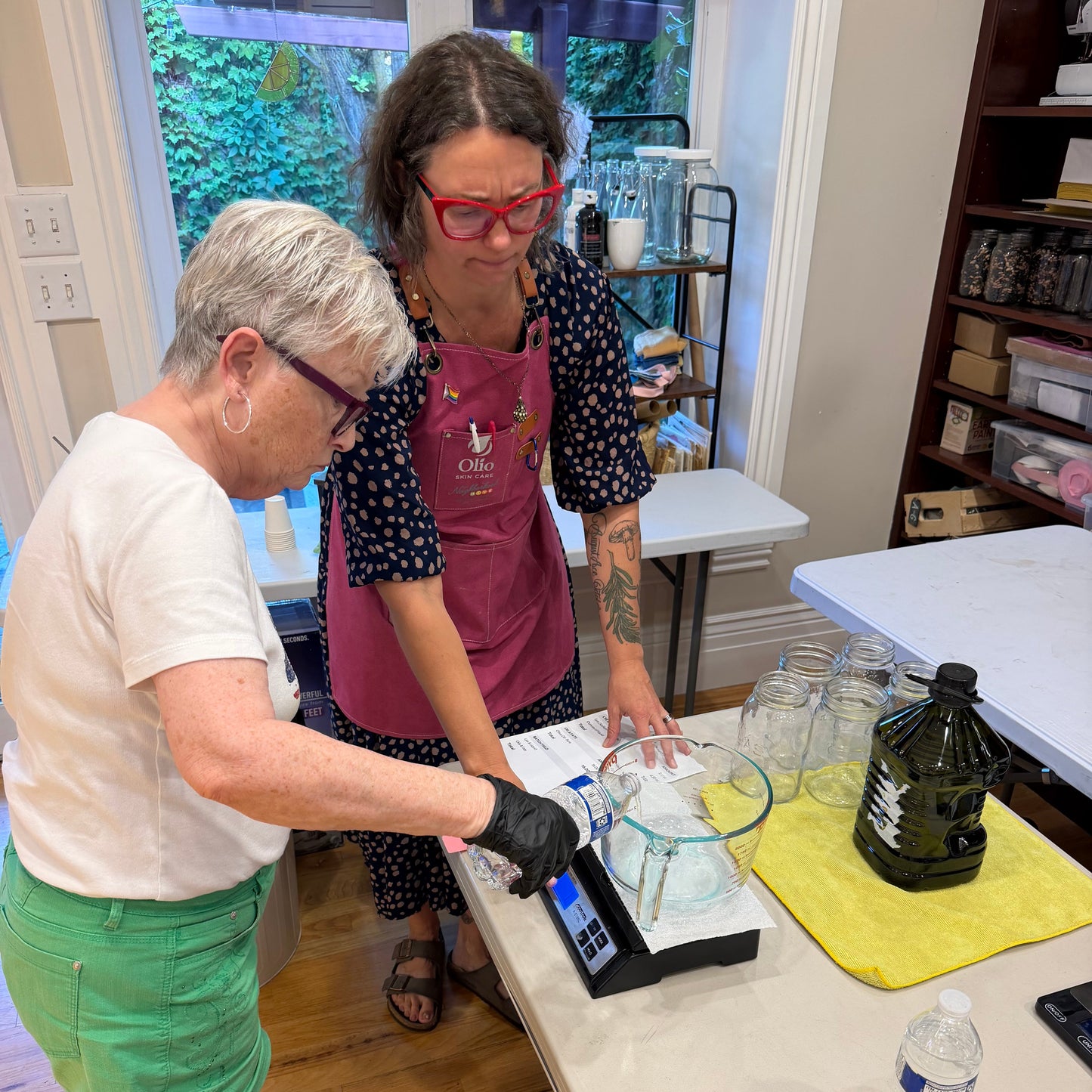 Two women standing at a table in a home kitchen or craft space, one weighing a clear glass bowl on a digital scale and the other observing. A yellow towel, a dark bottle, and other glassware are on the table.