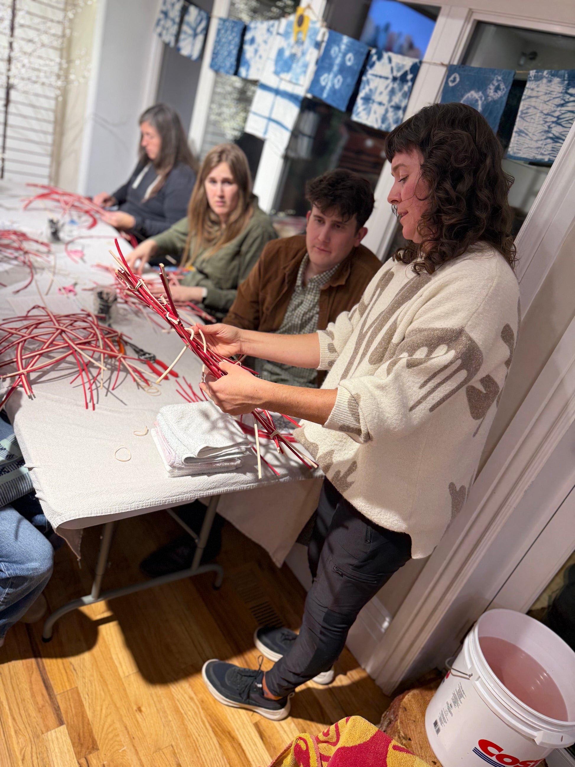 A group of people gathered around a table indoors, crafting with red and white materials. A woman in the foreground wears a cream knit sweater and dark pants, while others sit and work at the table.