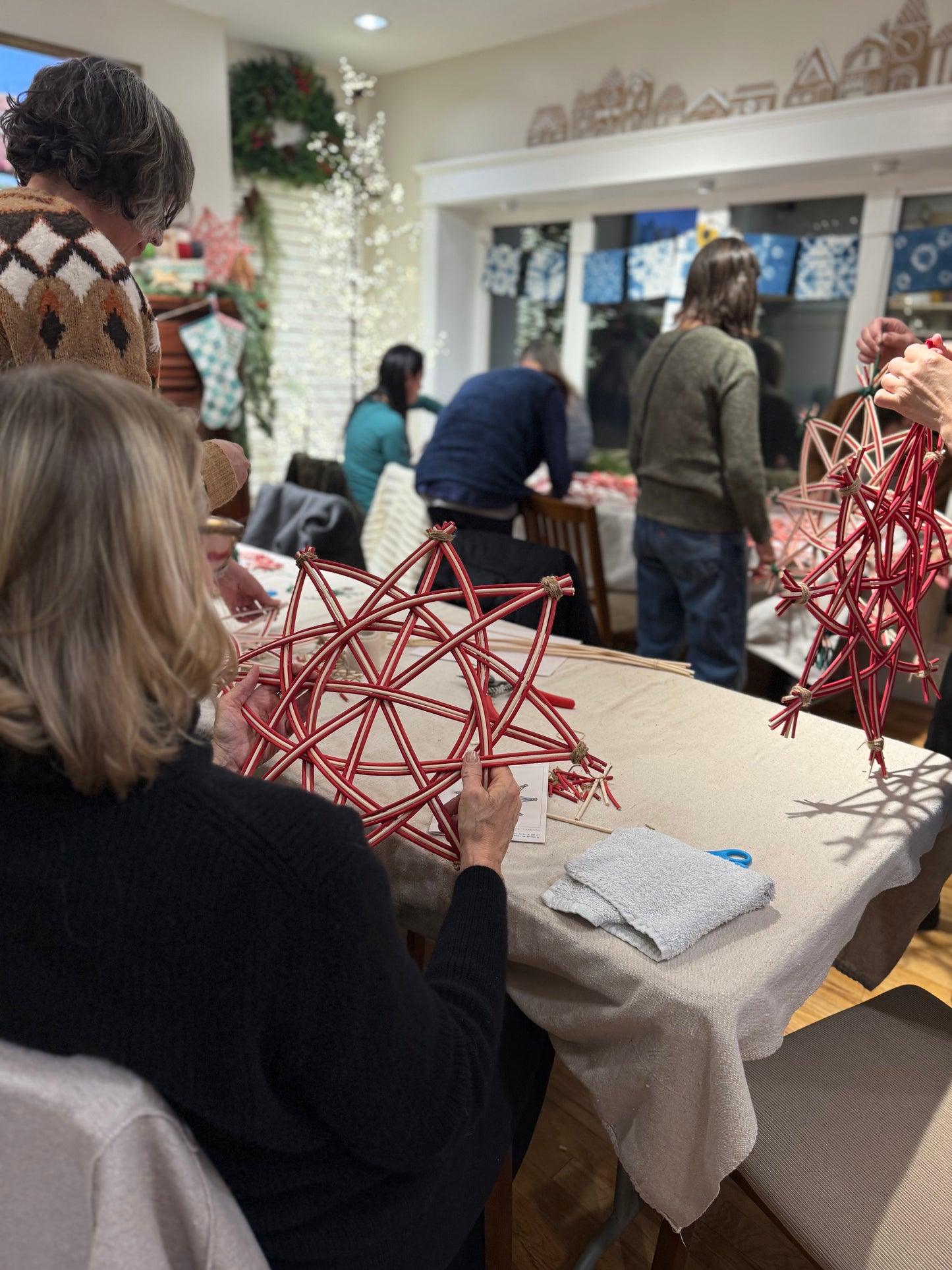 A group of people gathered around a table indoors, holding red geometric wire ornaments or decorative structures. The scene shows multiple people in a real indoor environment, so this is Lifestyle rather than Product/Packaging.