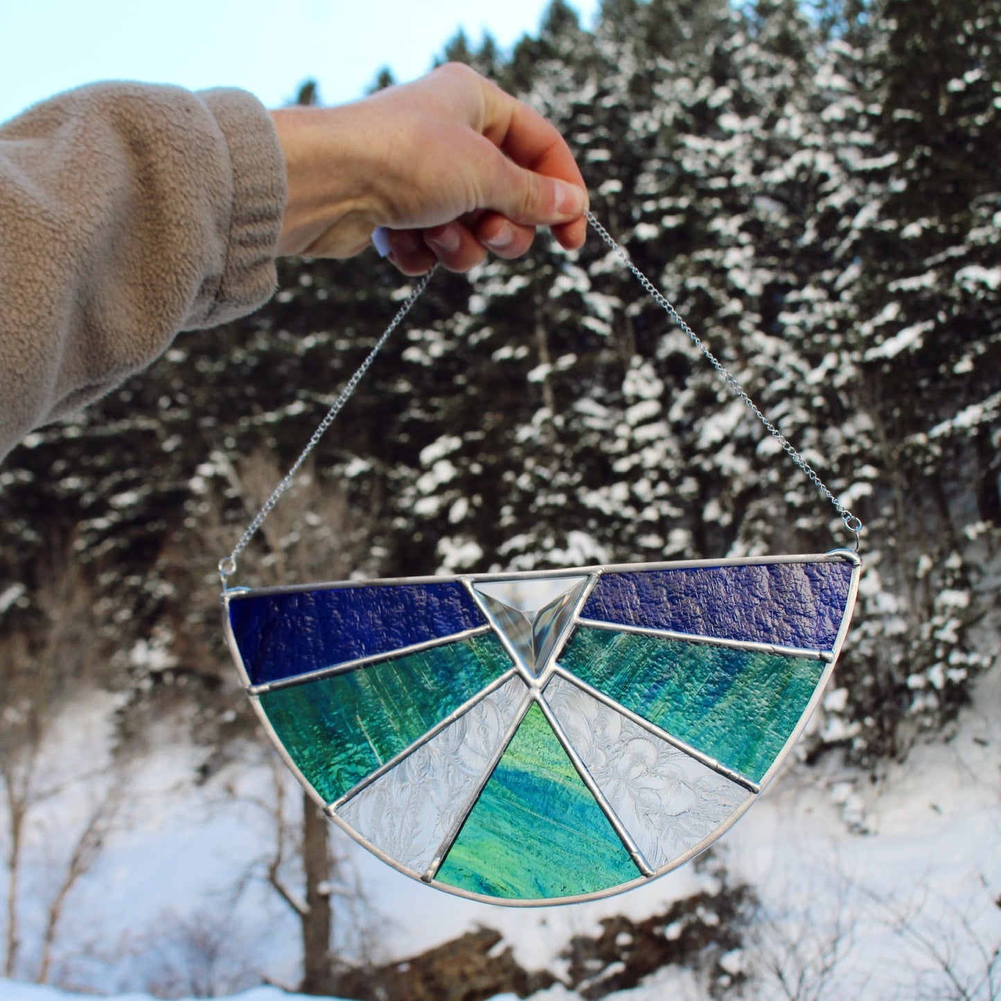 A hand holding a semi-circular stained glass suncatcher with blue, green, and white patterned glass panels, set against a snowy outdoor background with pine trees.